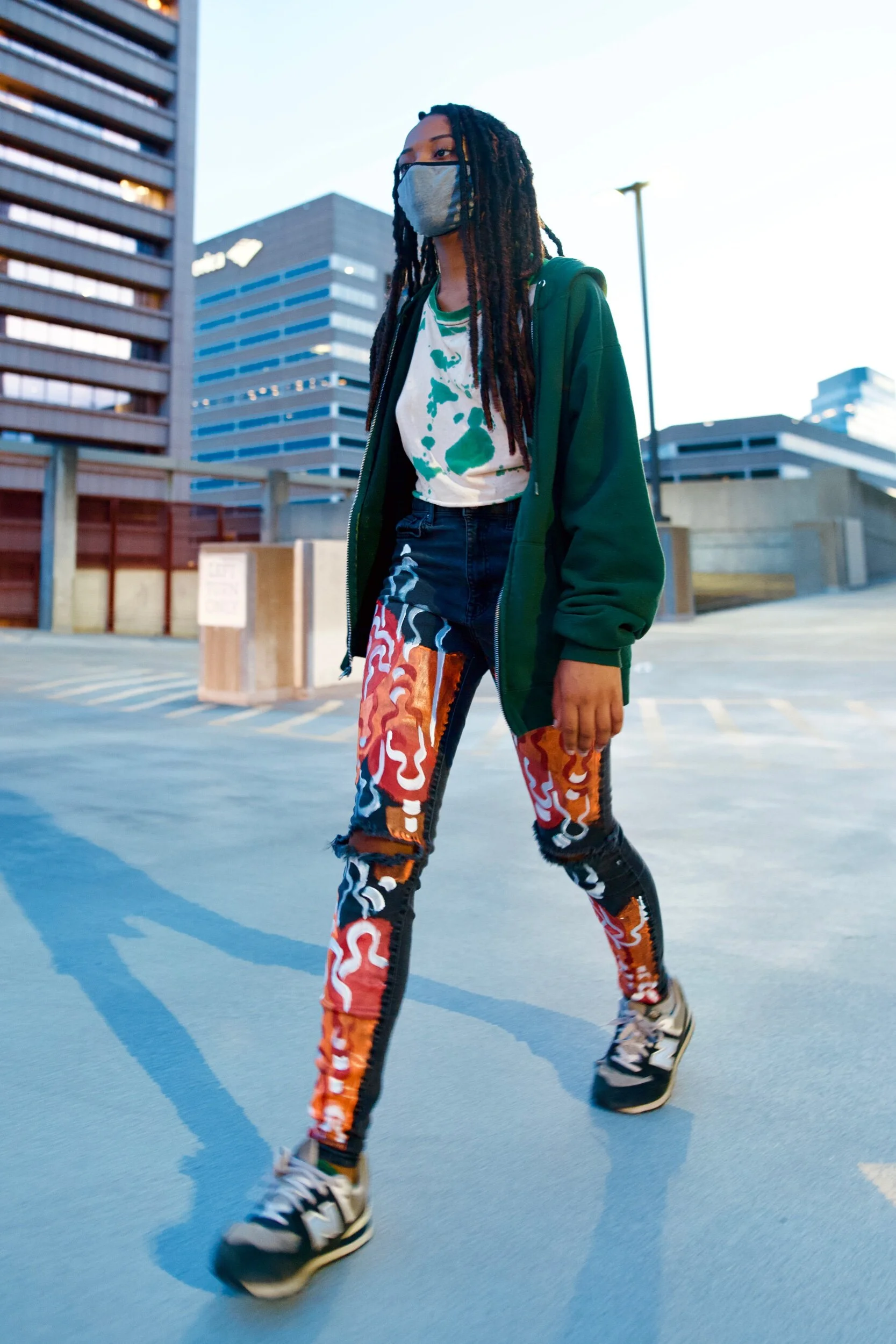 A young woman with long dreadlocks walking in an urban parking lot, wearing a face mask, green hoodie, tie-dye shirt, colorful patterned pants, and sneakers.