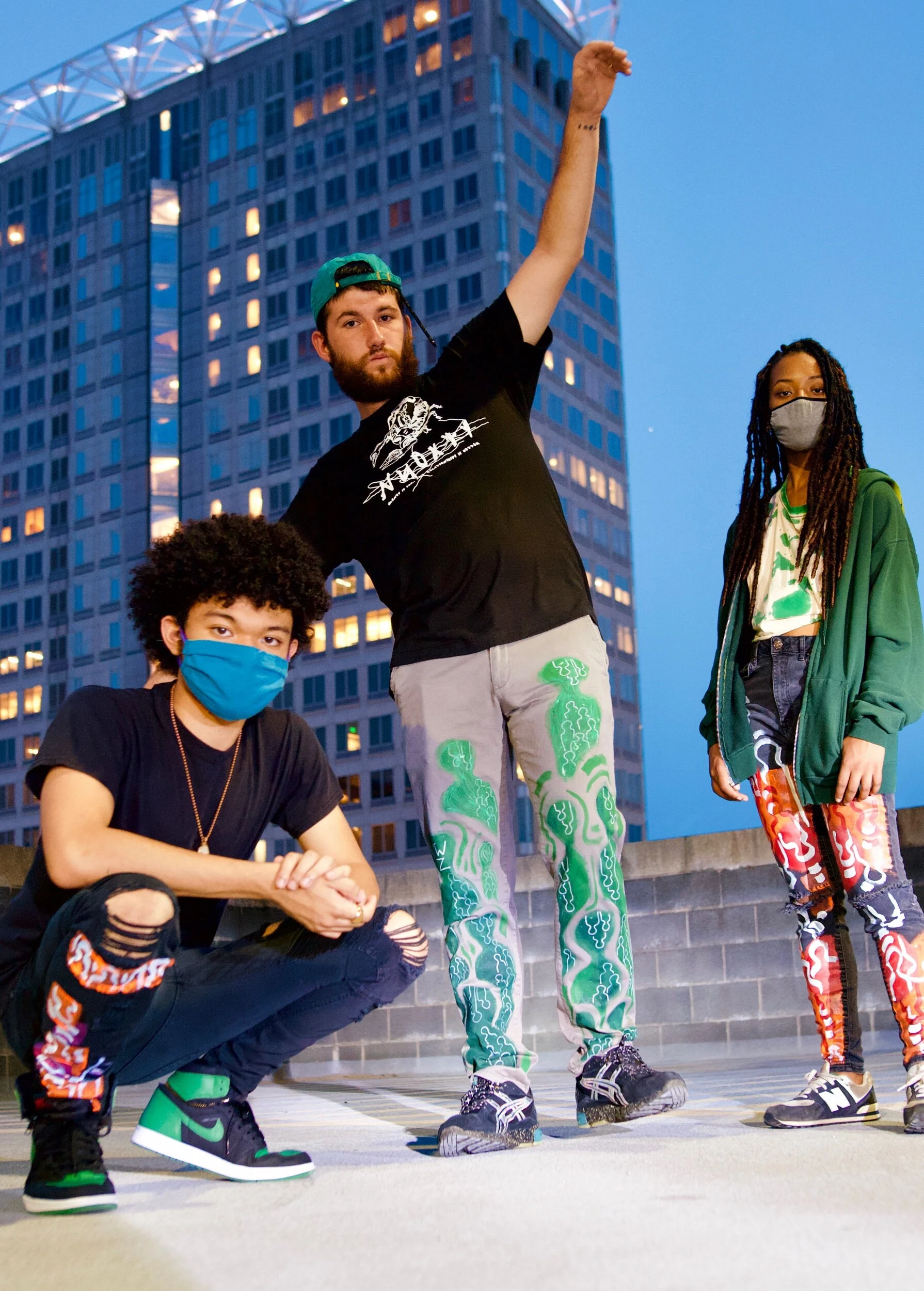 Three young people wearing masks, posing outdoors in front of a tall city building during dusk. One crouching, one standing with an arm raised, the third standing on the right side.