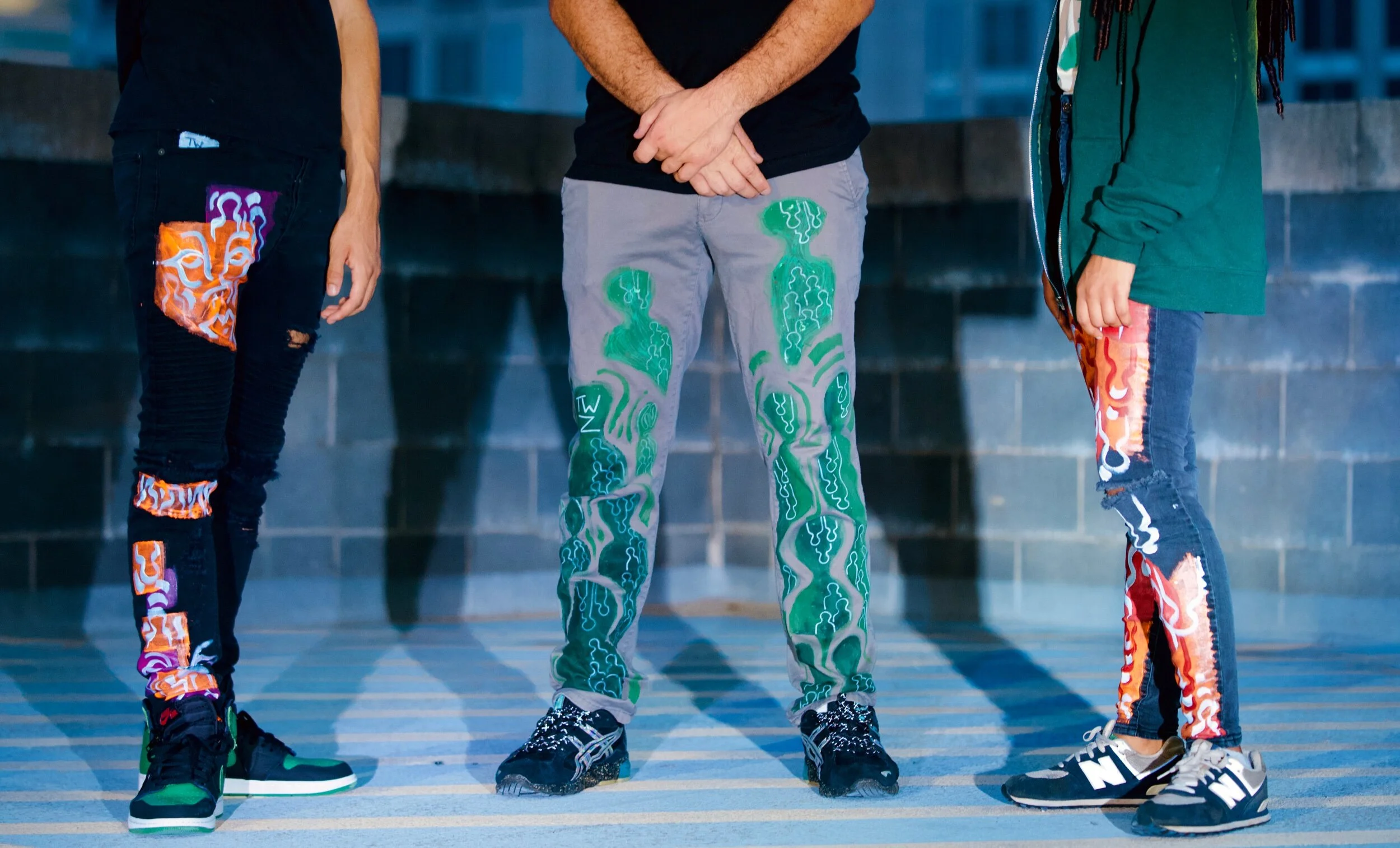 Close-up of three people standing outdoors, focusing on their legs and shoes, wearing colorful, patterned, distressed denim and pants with artistic designs.