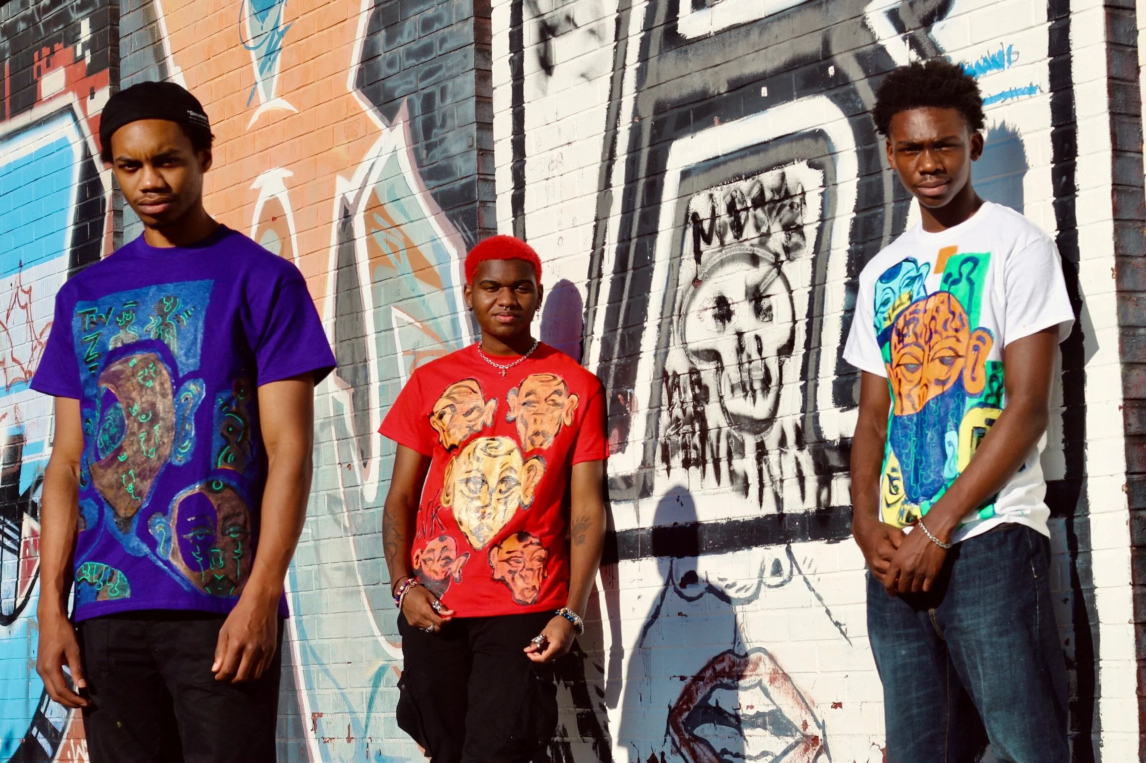 Three young men standing in front of a colorful graffiti mural on a brick wall, each wearing a vibrant shirt with artistic faces and designs.