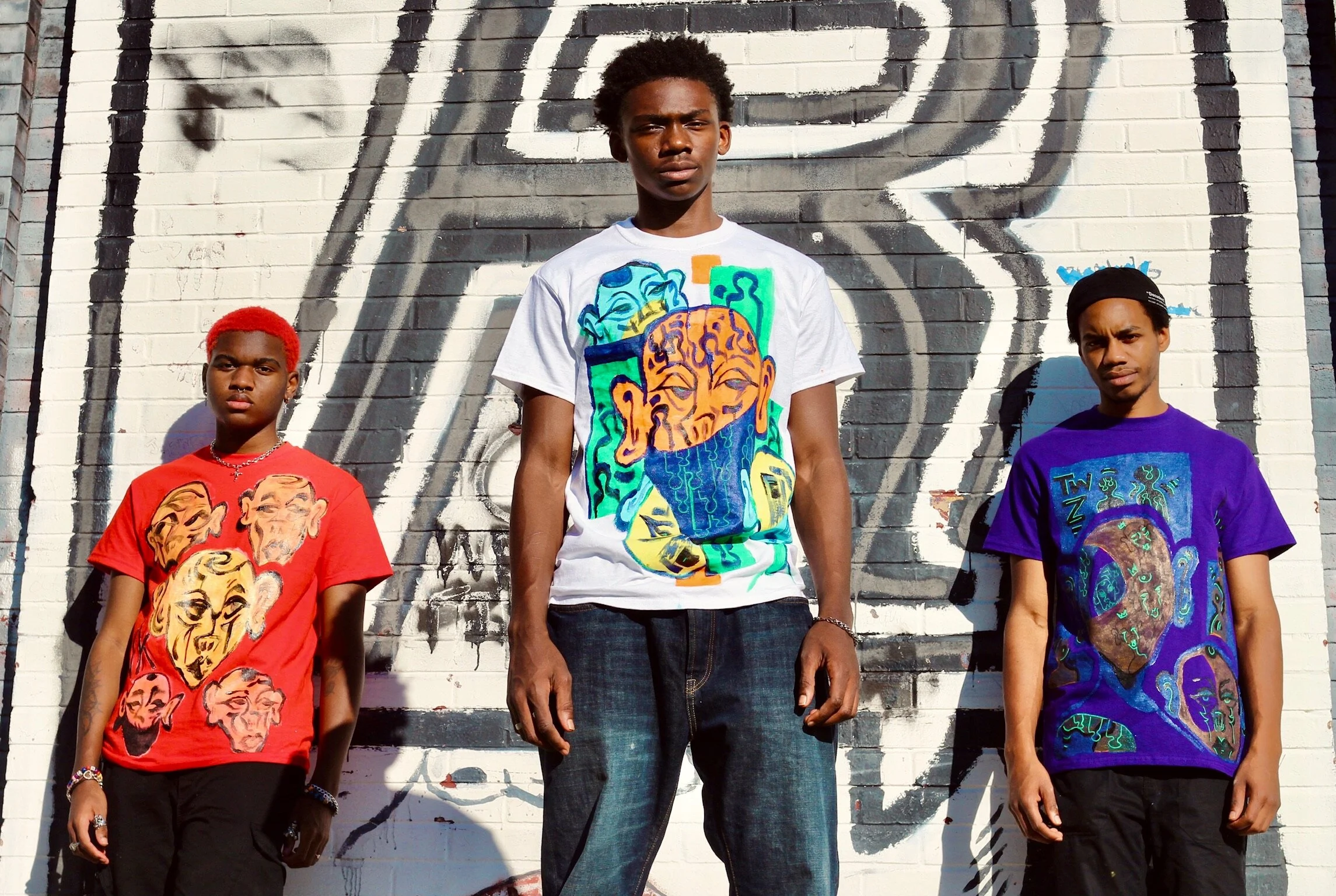 Three young men standing in front of a graffiti-covered brick wall, wearing colorful t-shirts with abstract face designs.