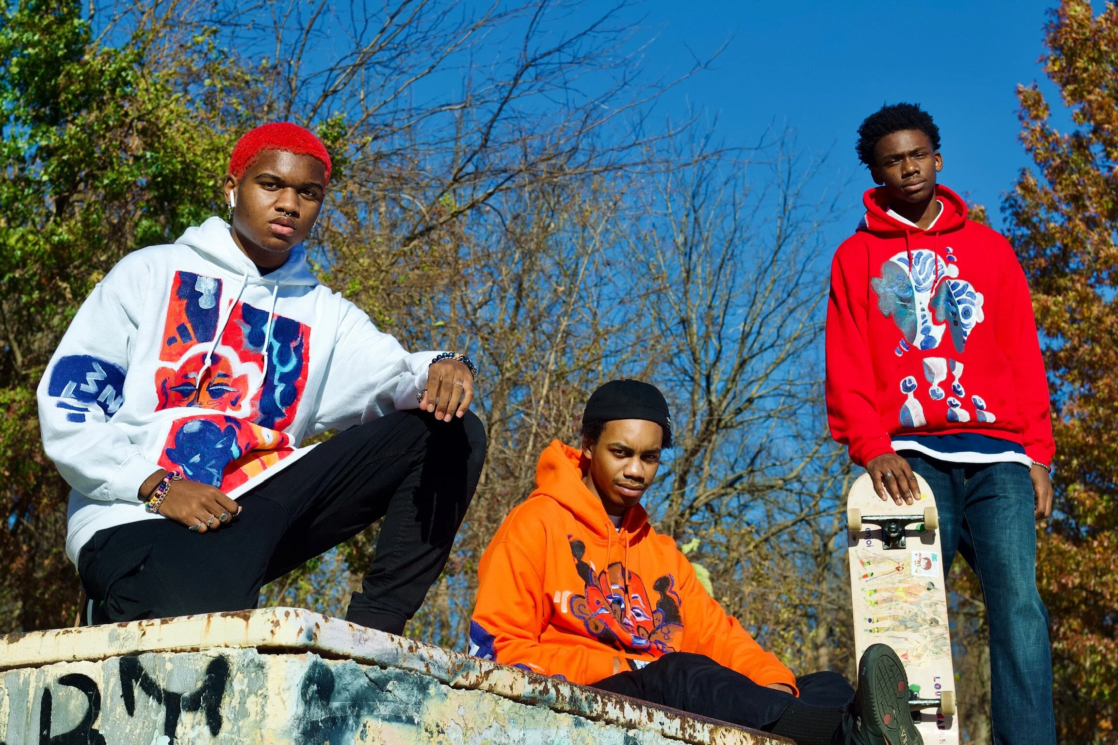 Three young men outdoors, two standing and one sitting with a skateboard, in front of trees with autumn leaves.