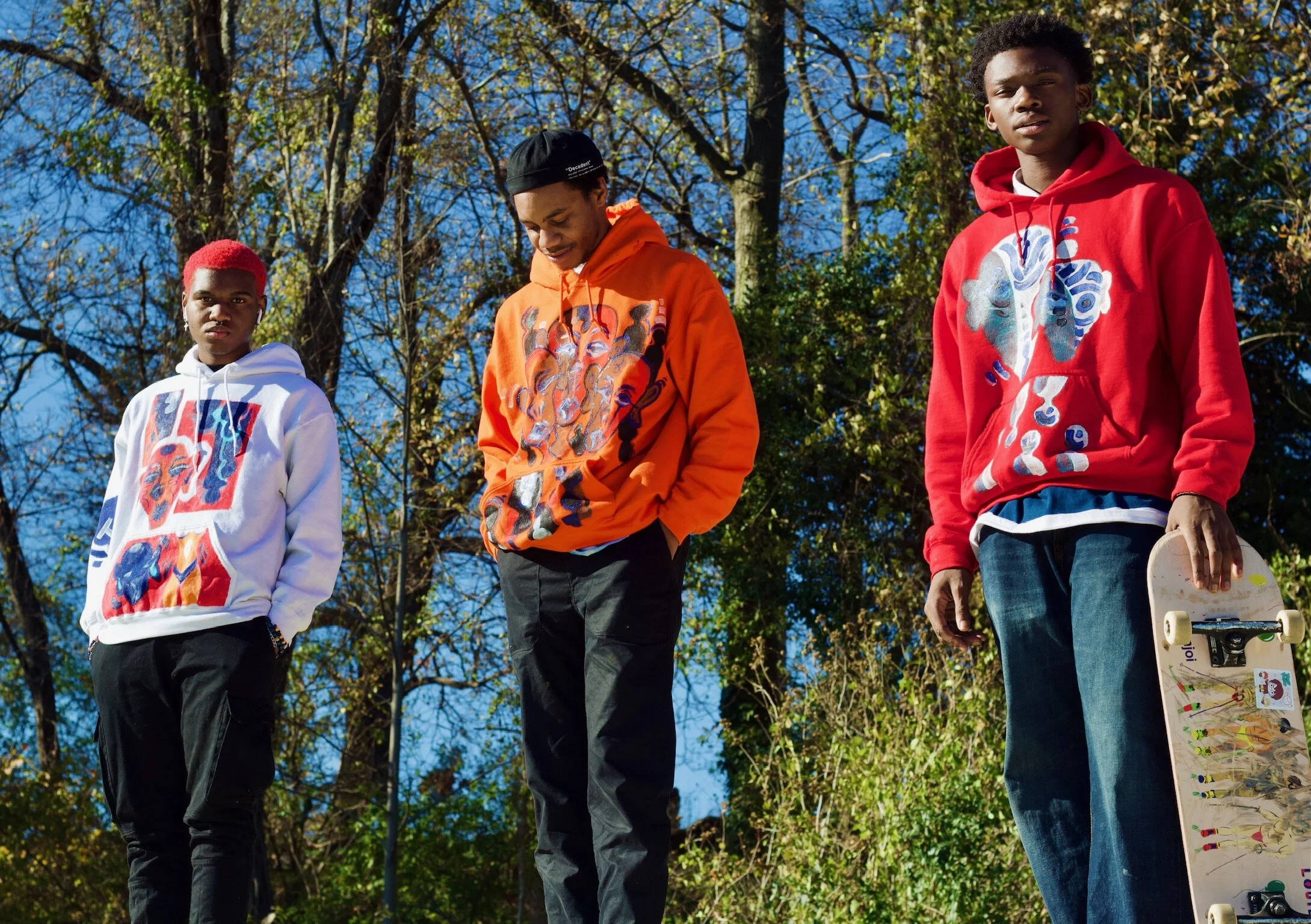 Three young men stand outdoors during daytime on a sunny autumn day, wearing colorful hoodies featuring artistic designs, with trees and blue sky in the background. One holds a skateboard.