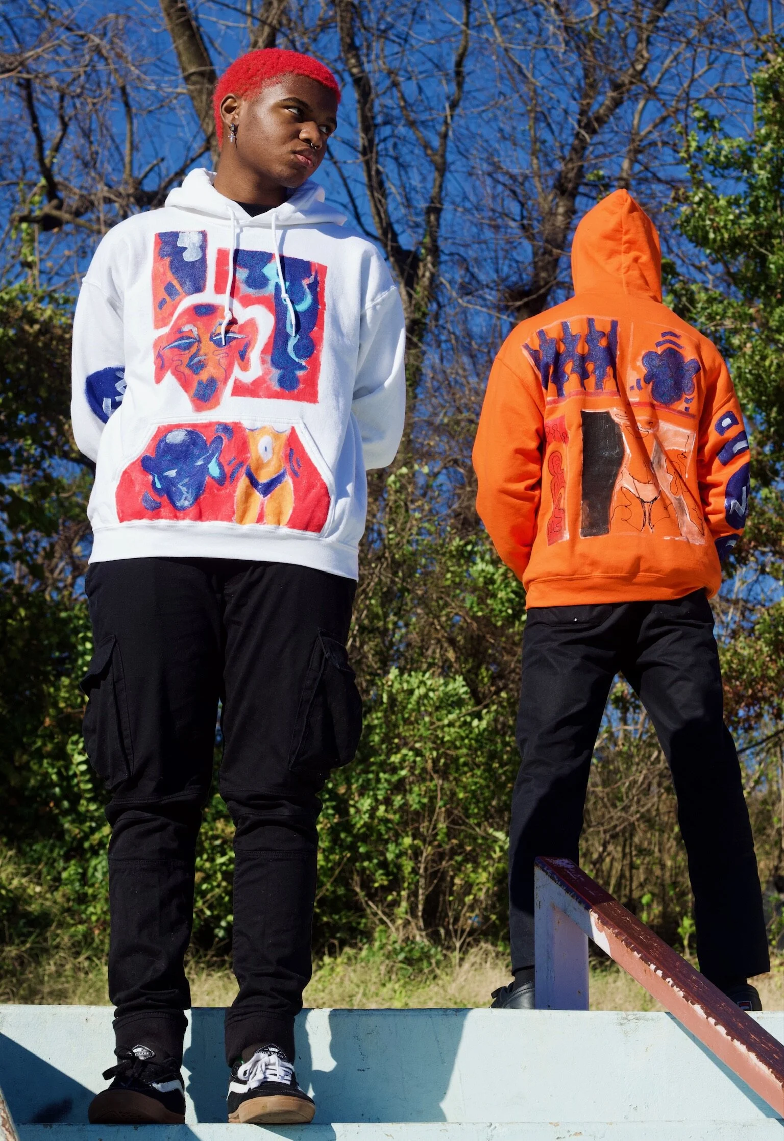 Two young men standing outdoors, one facing forward with red hair and wearing a white hoodie with colorful art designs, and the other with back turned wearing an orange hoodie with similar art designs.
