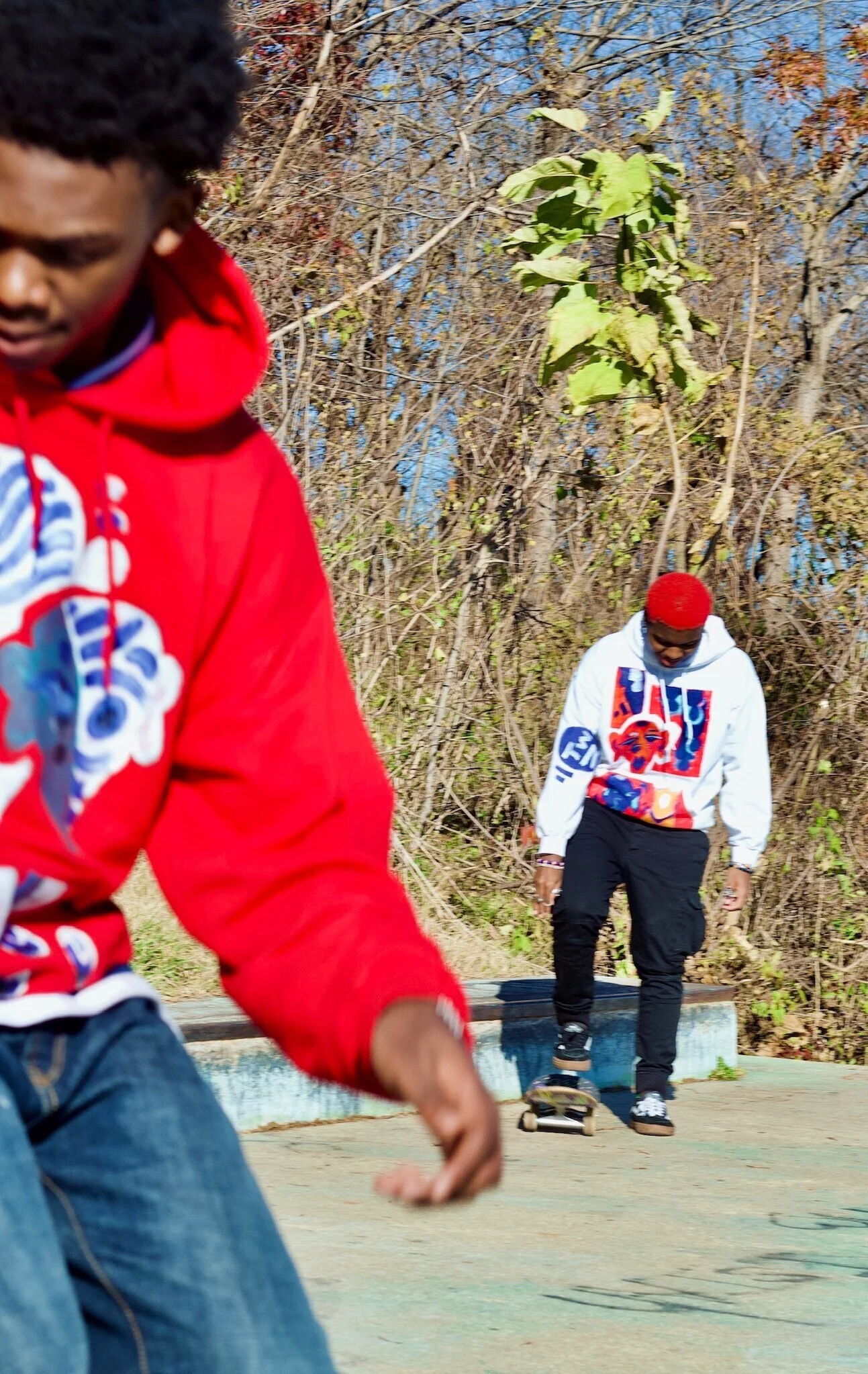 Two young men skateboarding outdoors on a sunny day, one in a white hoodie with colorful graphics and the other in a red hoodie.