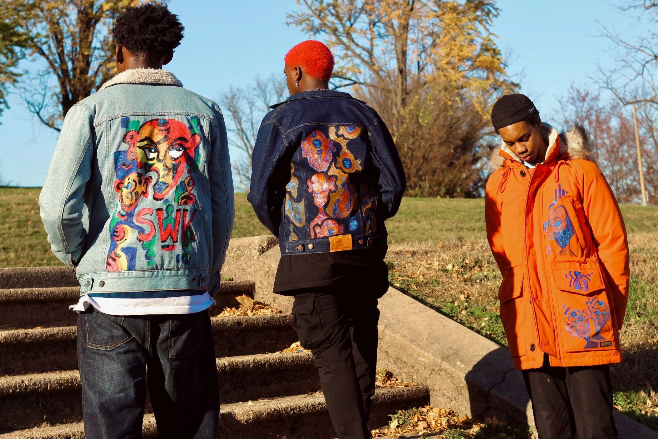 Three young men standing outdoors on a grassy area with fallen leaves, facing away from the camera. They are wearing colorful embroidered denim jackets with expressive designs, one in light wash, one in dark wash, and one in bright orange.