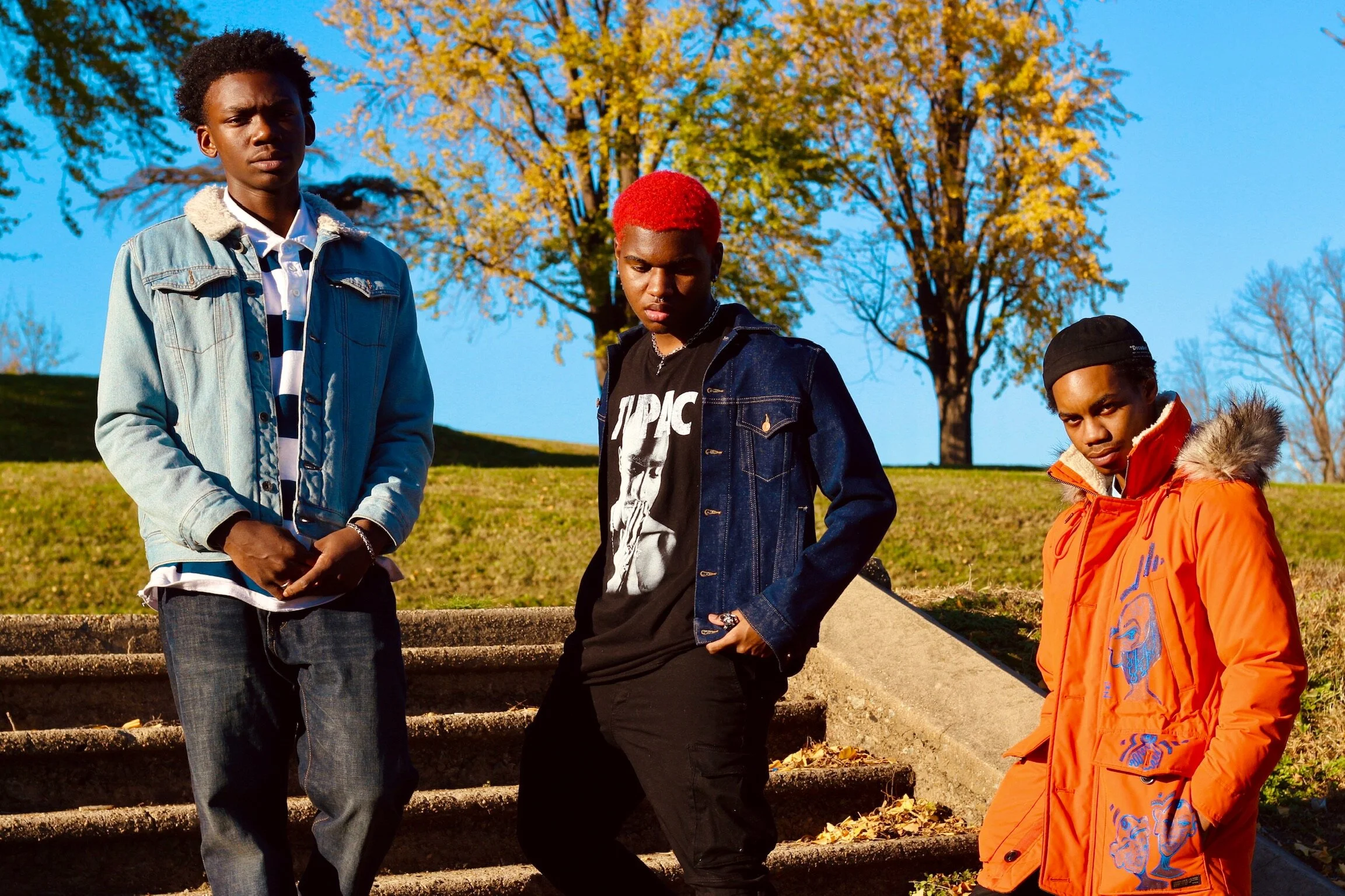 Three young men standing on outdoor stairs during autumn, with trees and a bright blue sky in background. One wearing a denim jacket, one with red hair and a black T-shirt, and another in an orange jacket with a fur-lined hood.
