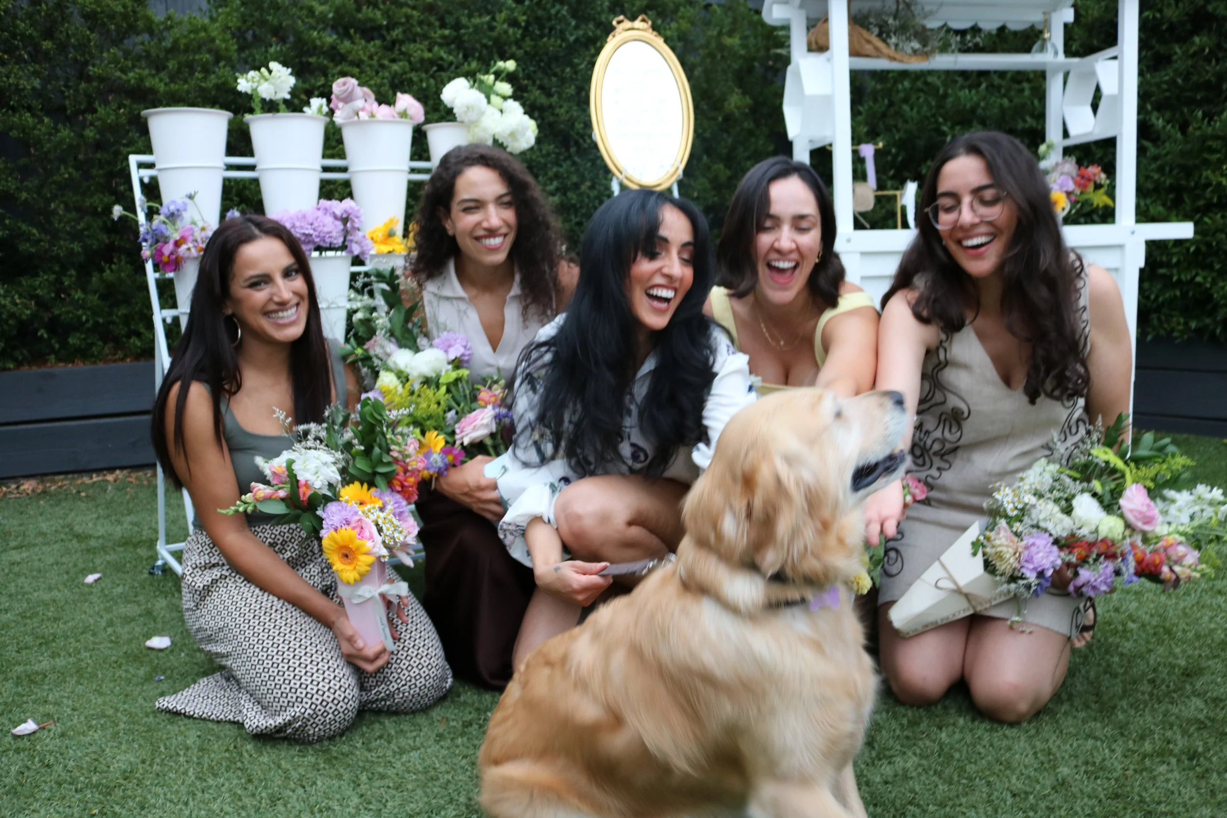 Five women sitting on the grass at a garden party, laughing and smiling, with a large golden retriever dog in front of them. They hold bouquets of flowers and are surrounded by floral arrangements and greenery.