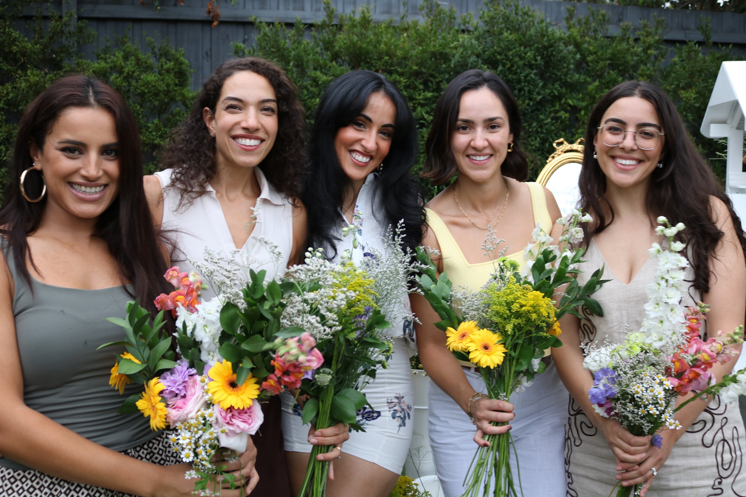 Five women smiling and holding bouquets of colorful flowers in an outdoor setting.