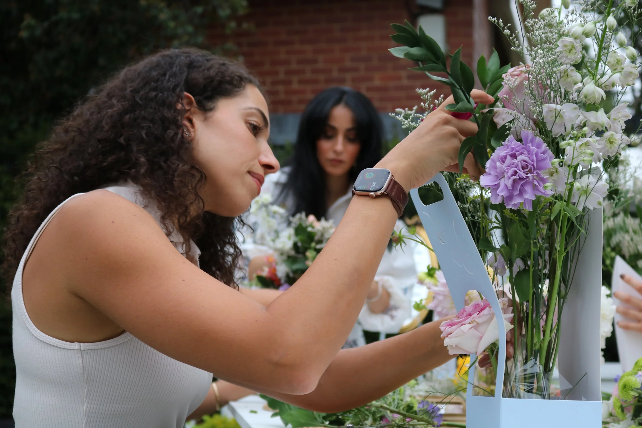 A woman with curly hair arranging flowers in a white container during a garden event, with another woman visible in the background.