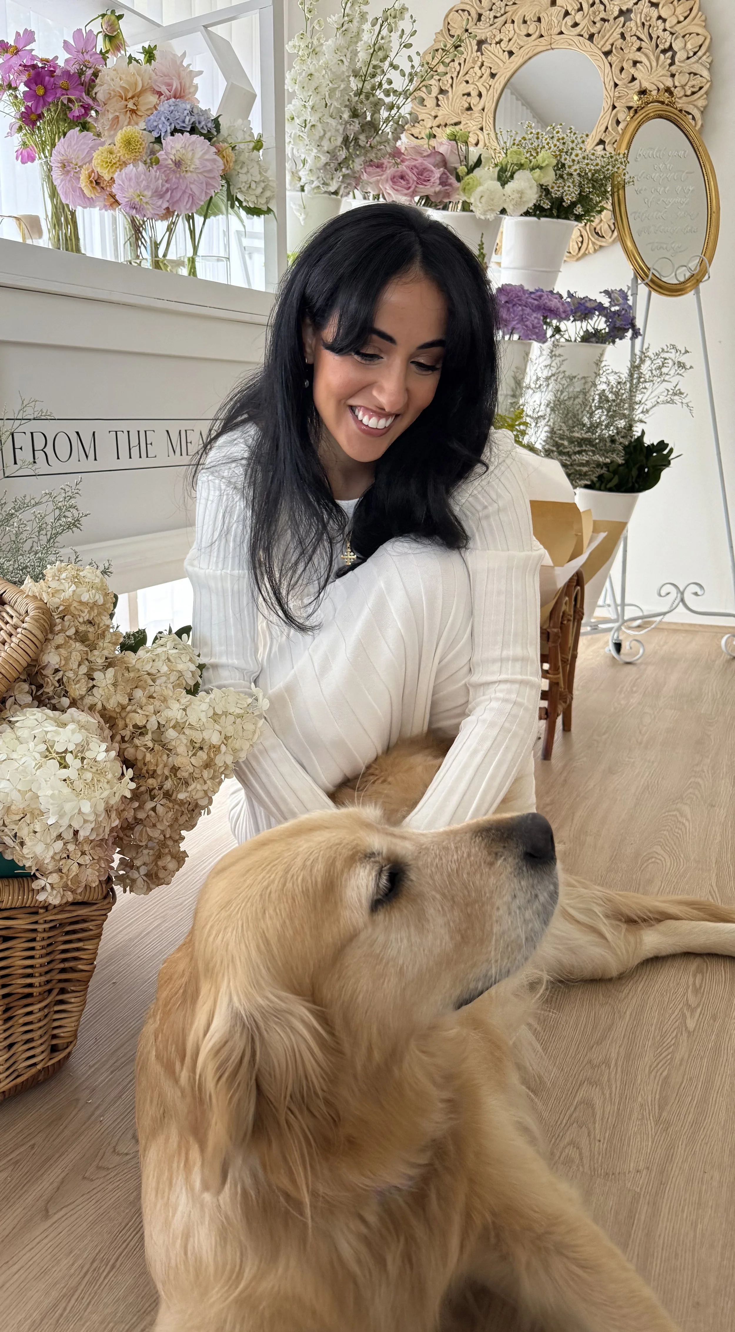 A woman with dark hair smiling while sitting on the floor, next to a golden retriever dog resting with eyes closed, surrounded by a variety of colorful flowers and decorative mirrors in a bright, floral indoor setting.