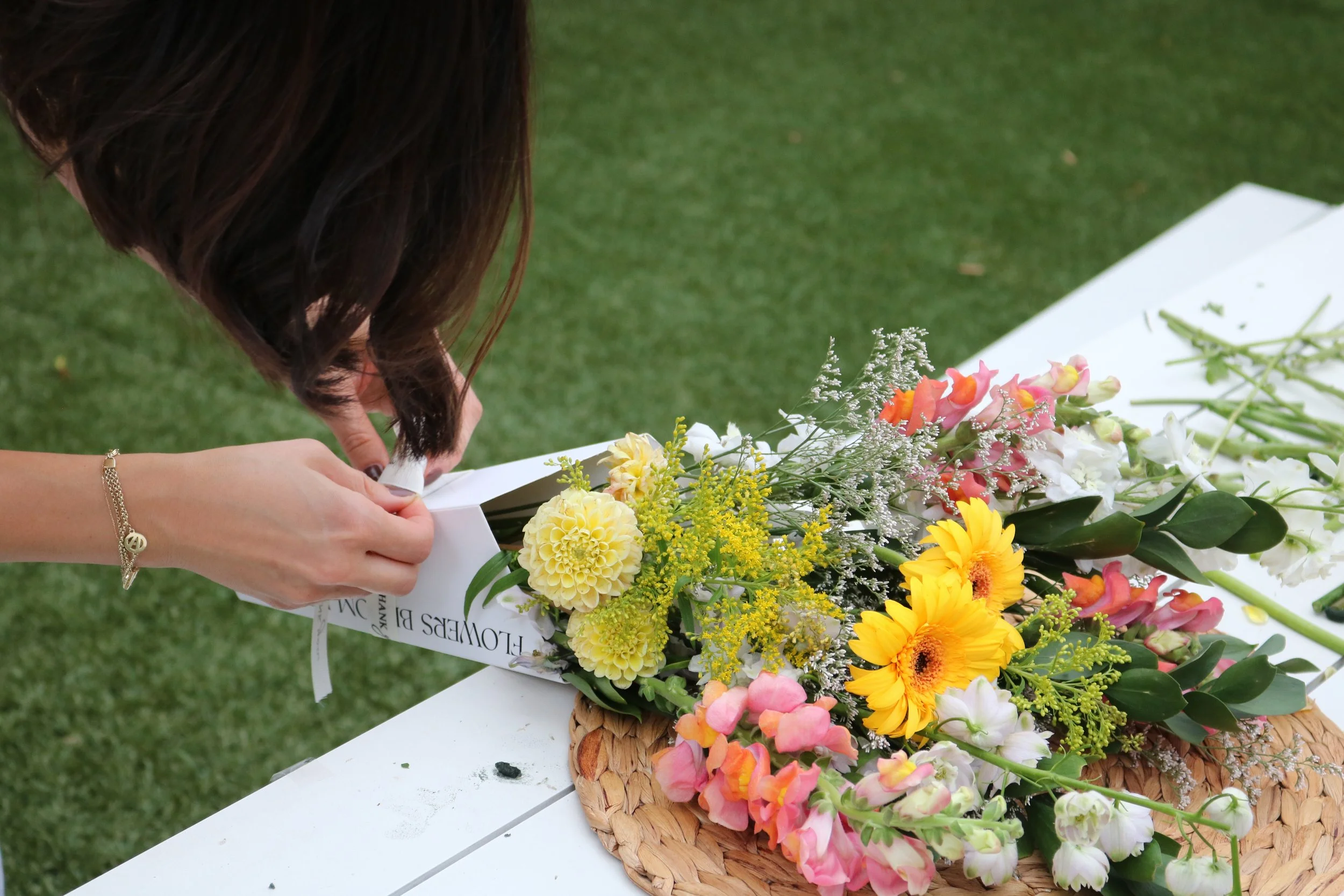 Woman arranging a colorful flower bouquet on a white table outdoors.