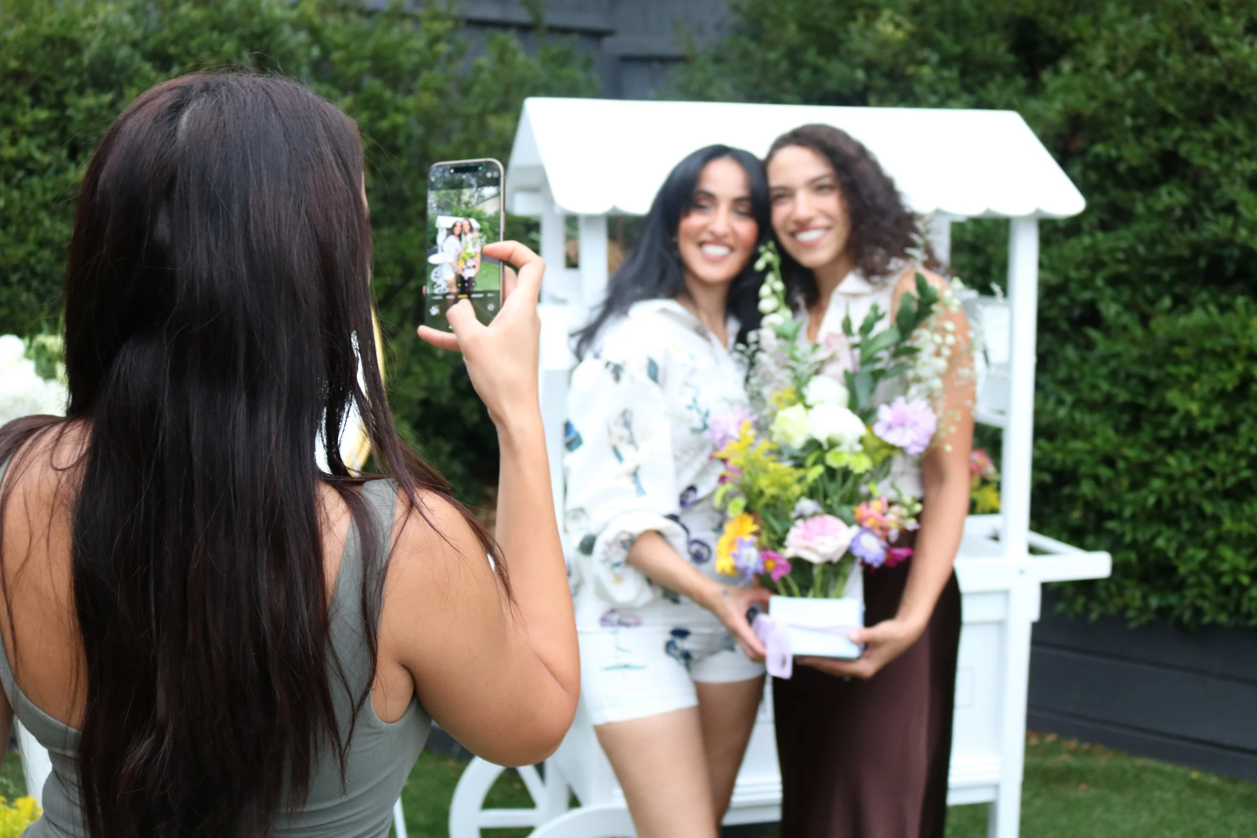 A woman takes a photo of two women with a mobile phone at an outdoor garden setting, with one holding a large bouquet of flowers and both smiling.