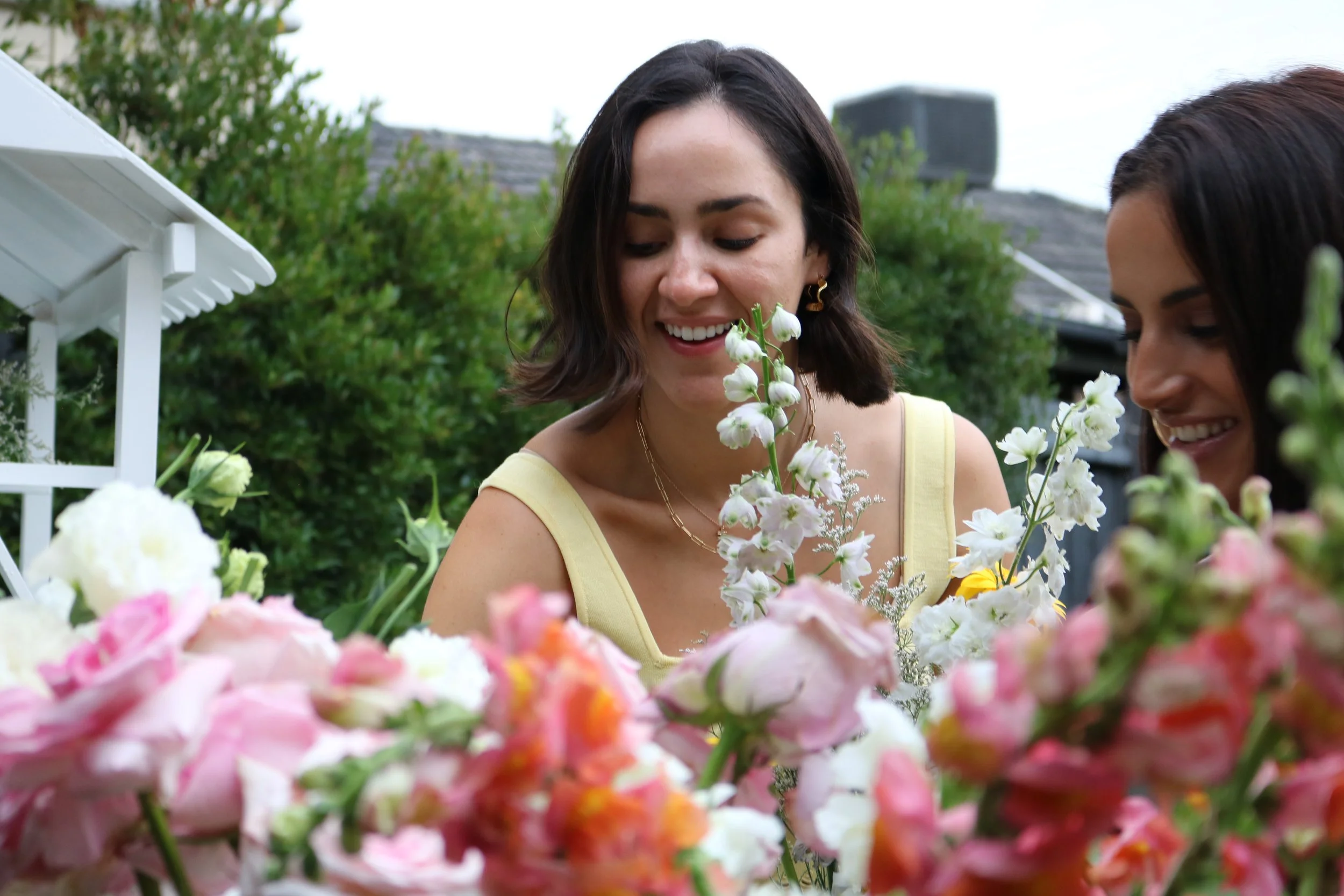 Two women smiling and looking at colorful flowers in a garden.