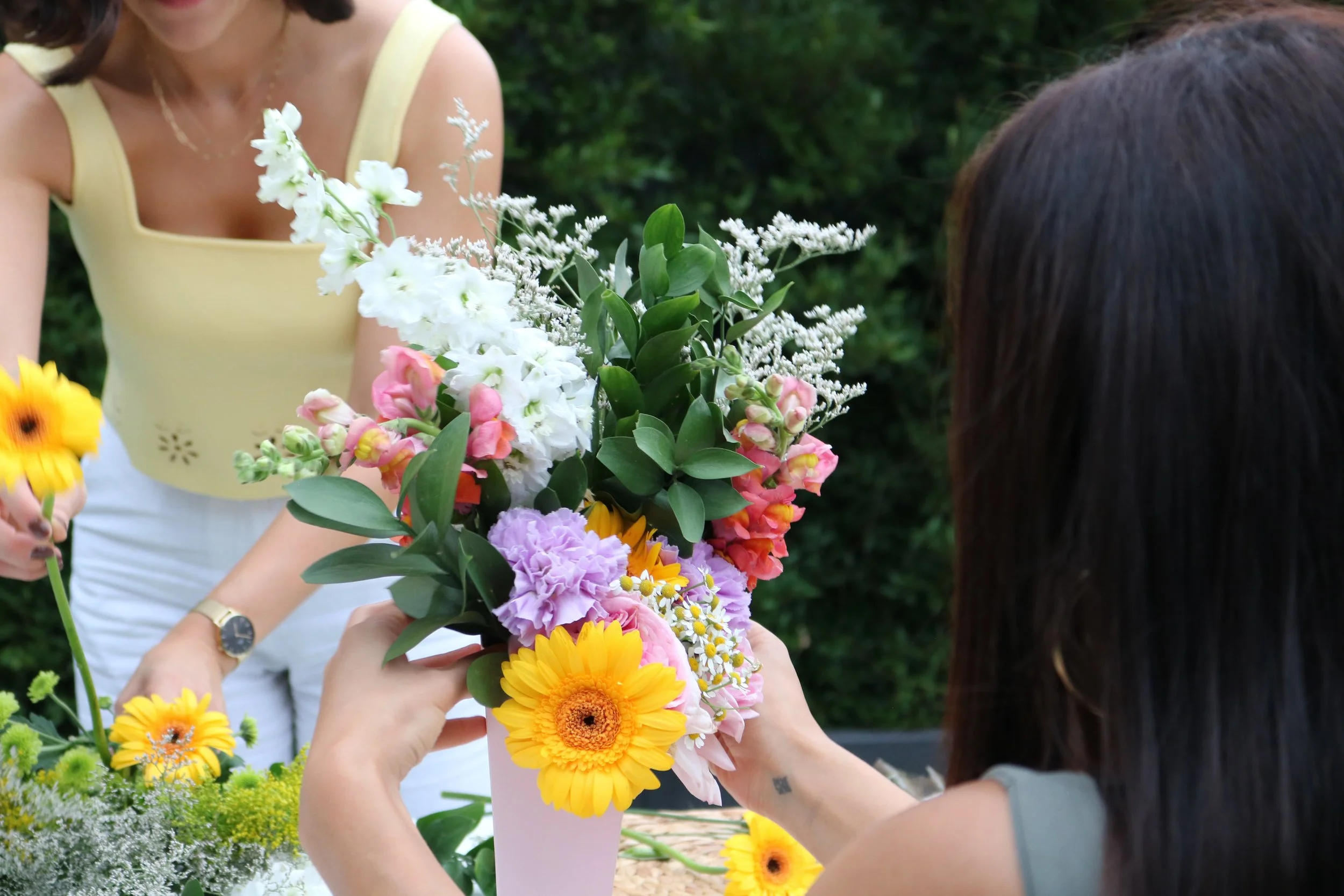A woman with dark hair arranging a colorful bouquet of flowers, including yellow, pink, purple, and white blooms, outdoors with greenery in the background.