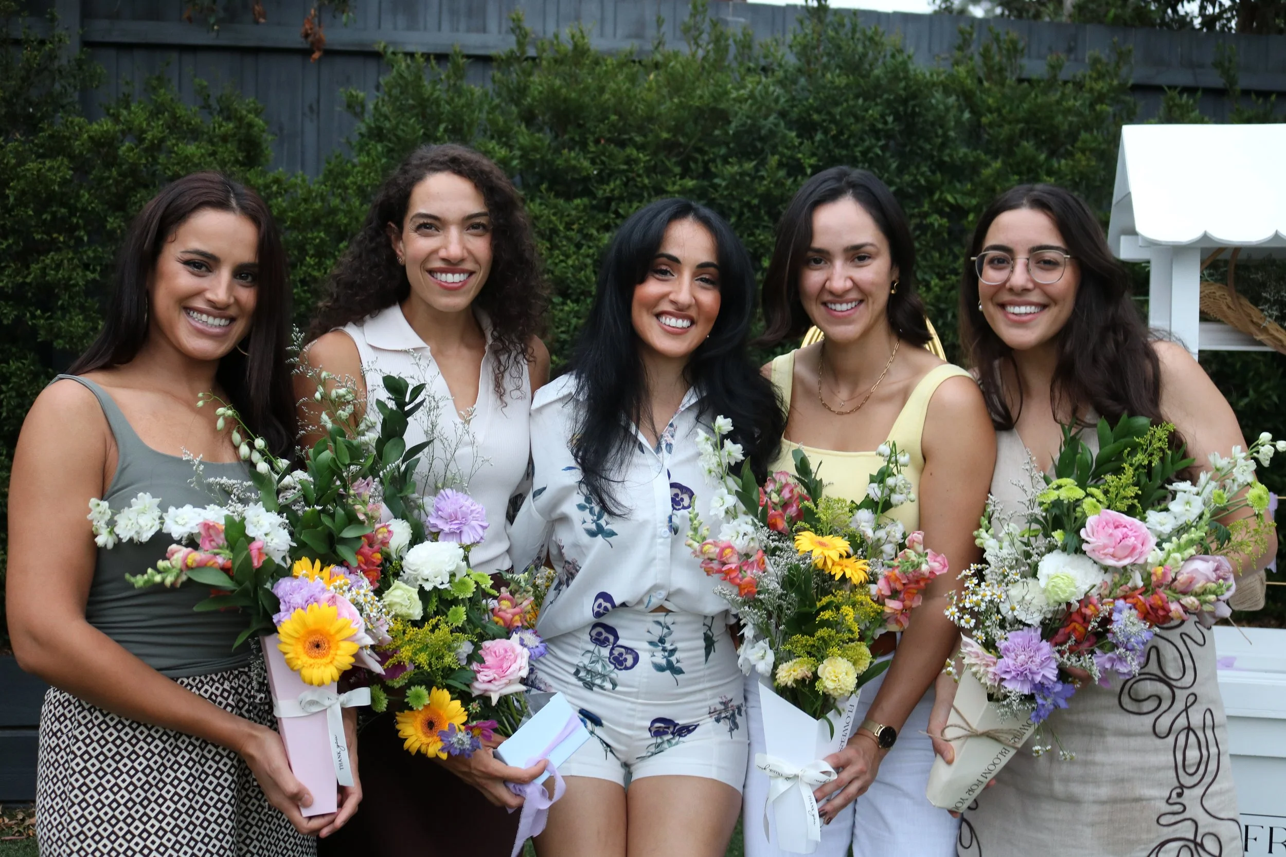 Five women smiling and holding bouquets of colorful flowers outdoors, with greenery and a fence in the background.