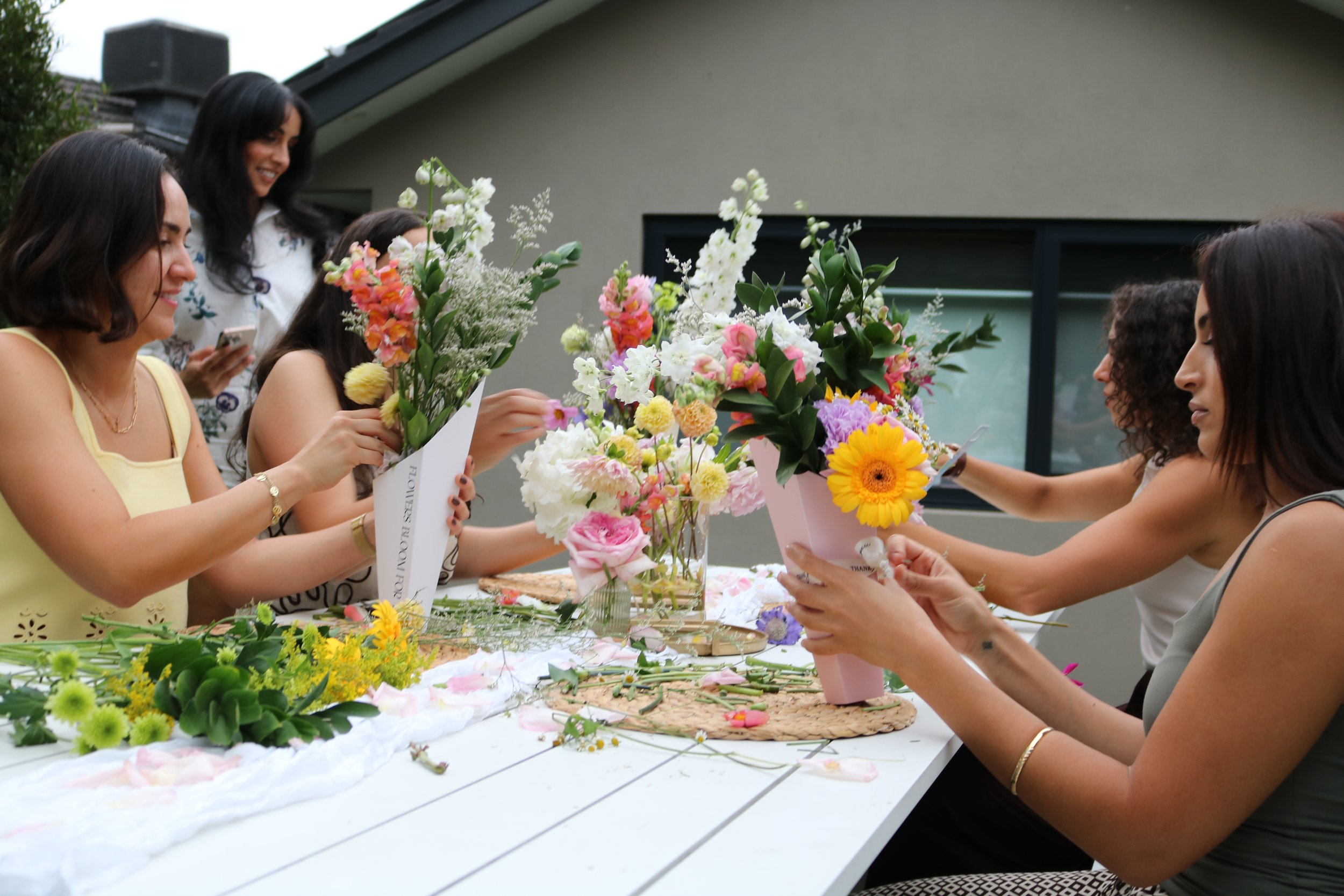 A group of women are making and arranging floral bouquets on a table outdoors.