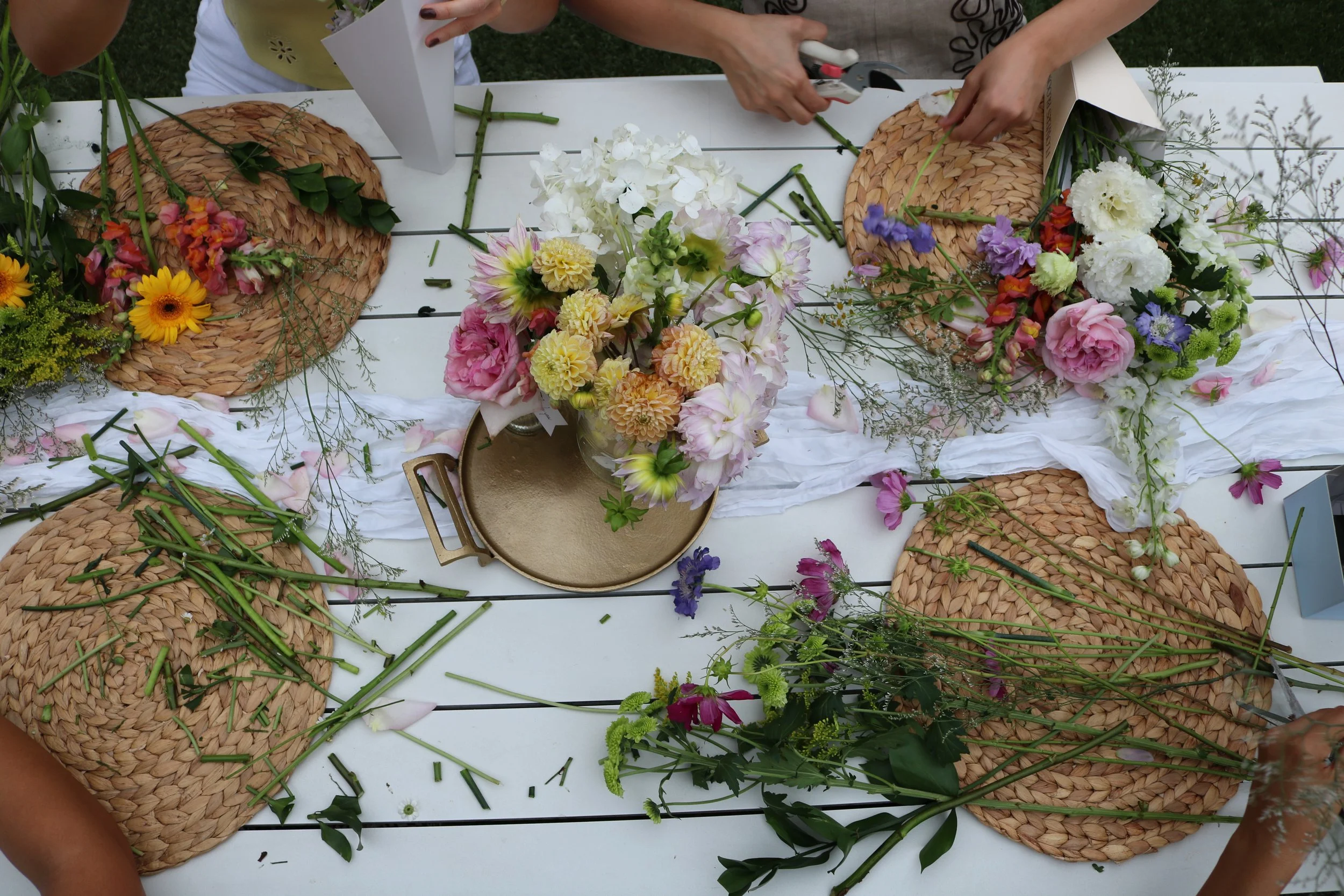 People arranging colorful flower bouquets on a white table with woven placemats and white cloth runners, preparing for a floral arrangement event.