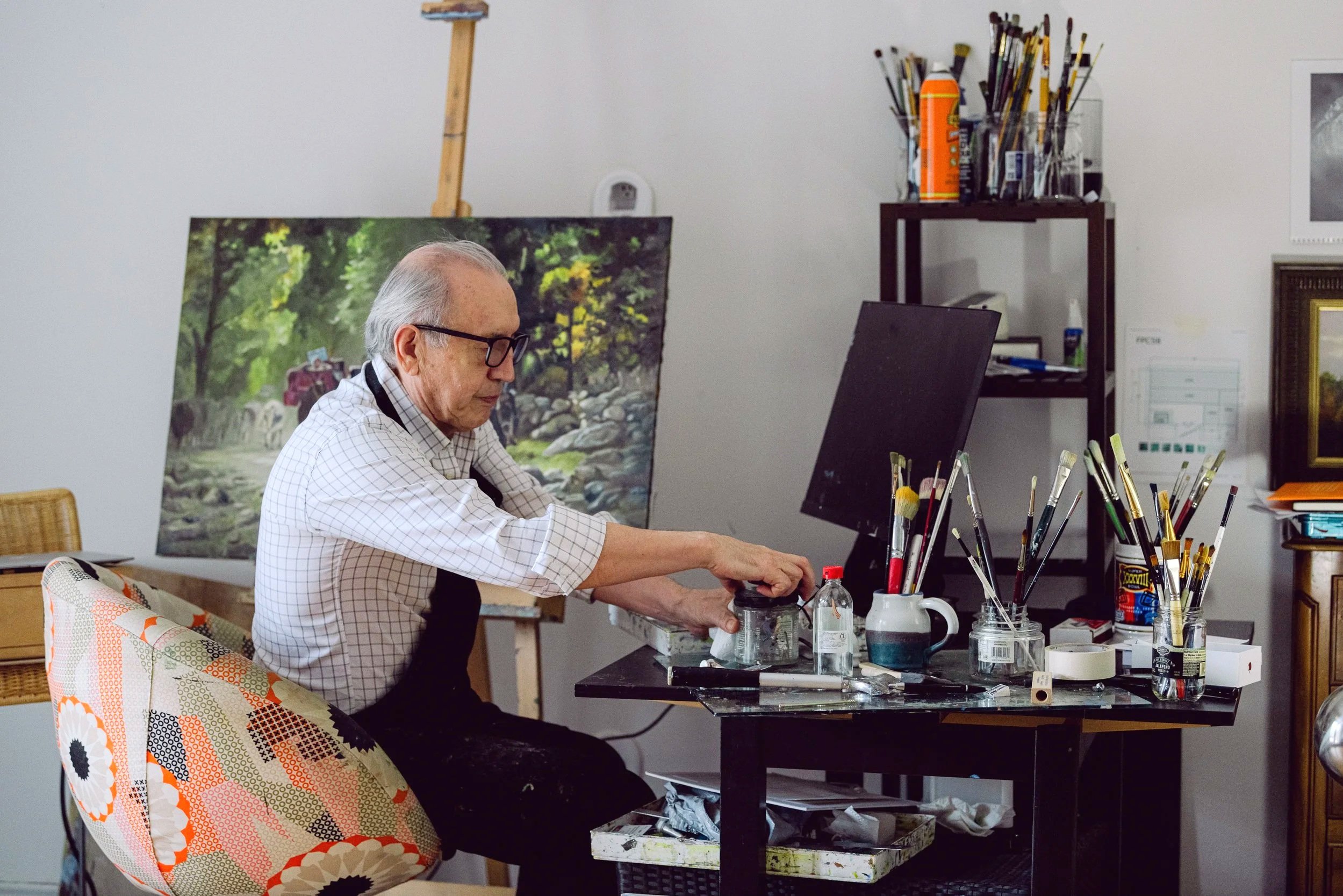 An elderly man with gray hair and glasses working at an art studio desk, surrounded by paintbrushes, art supplies, and artwork, including a landscape painting and a computer monitor.