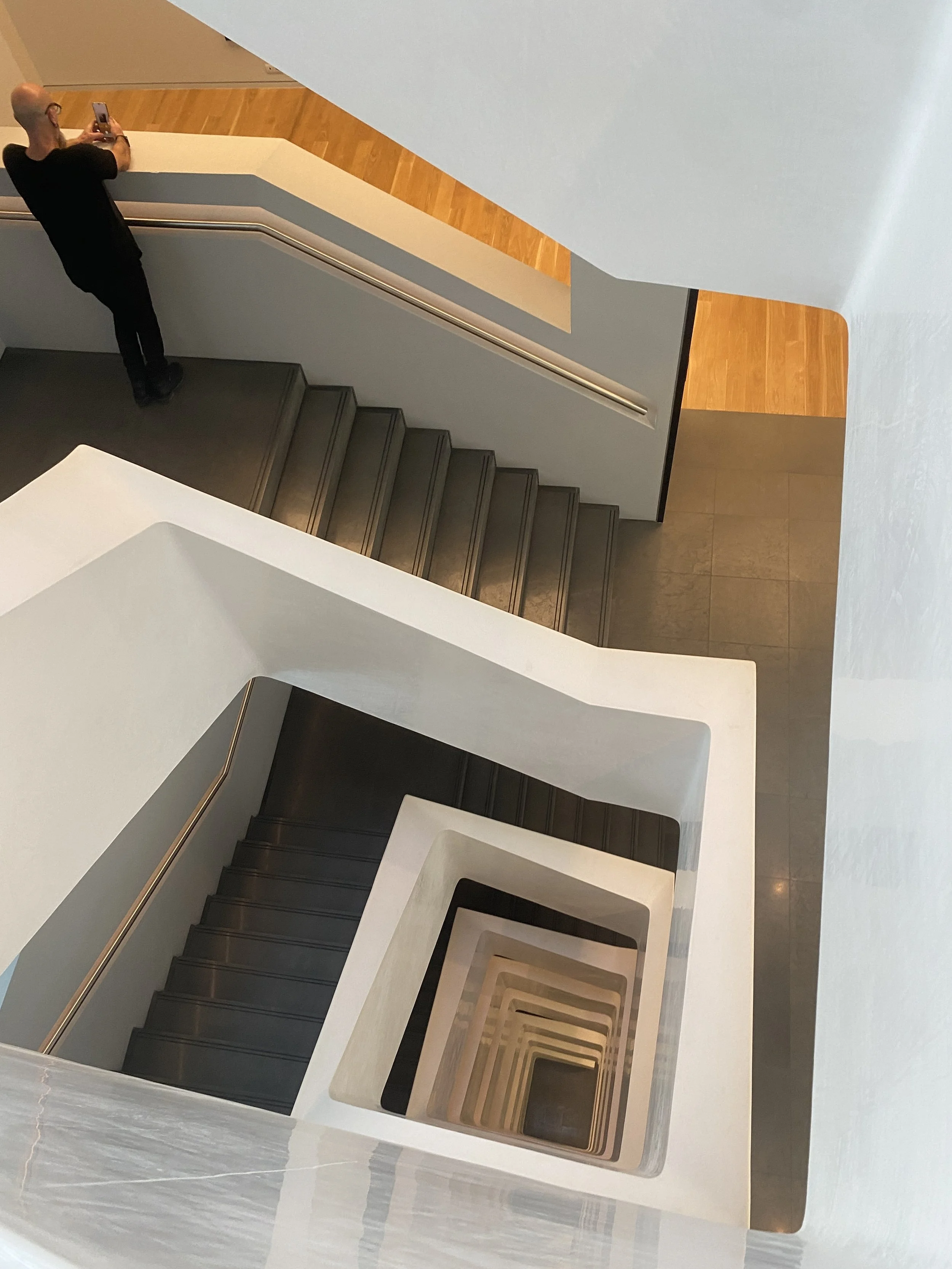 Looking down a spiral staircase with steps in shades of black and beige, viewed from the top, with a person taking a photo at the top.