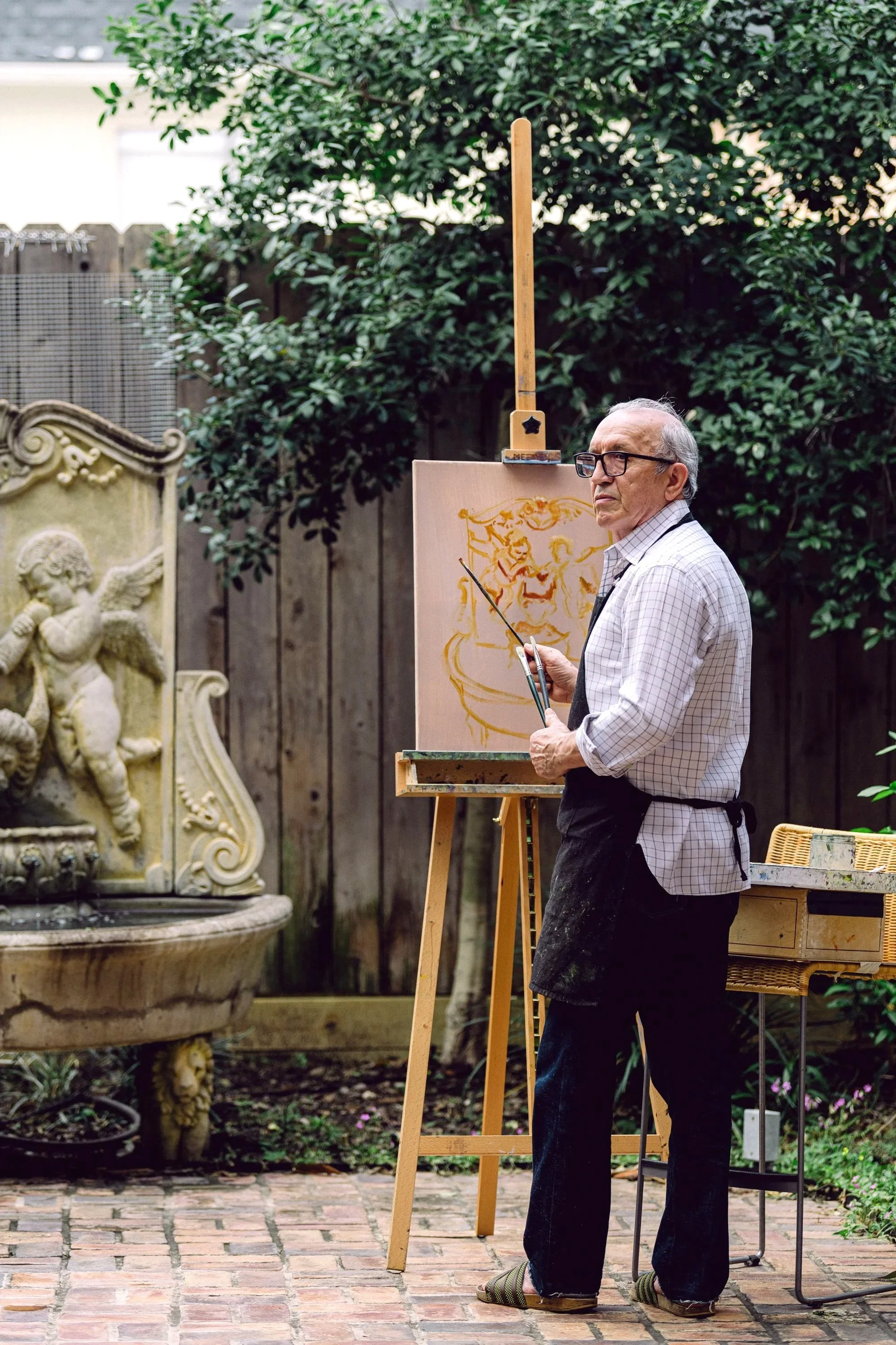 An elderly man with glasses and gray hair, wearing a checkered shirt, black apron, dark pants, and striped slippers, is painting a yellow design on a canvas outdoors. He holds a paintbrush, standing in a backyard with a wooden fence, green trees, a stone fountain with a cherub, and gardening equipment.