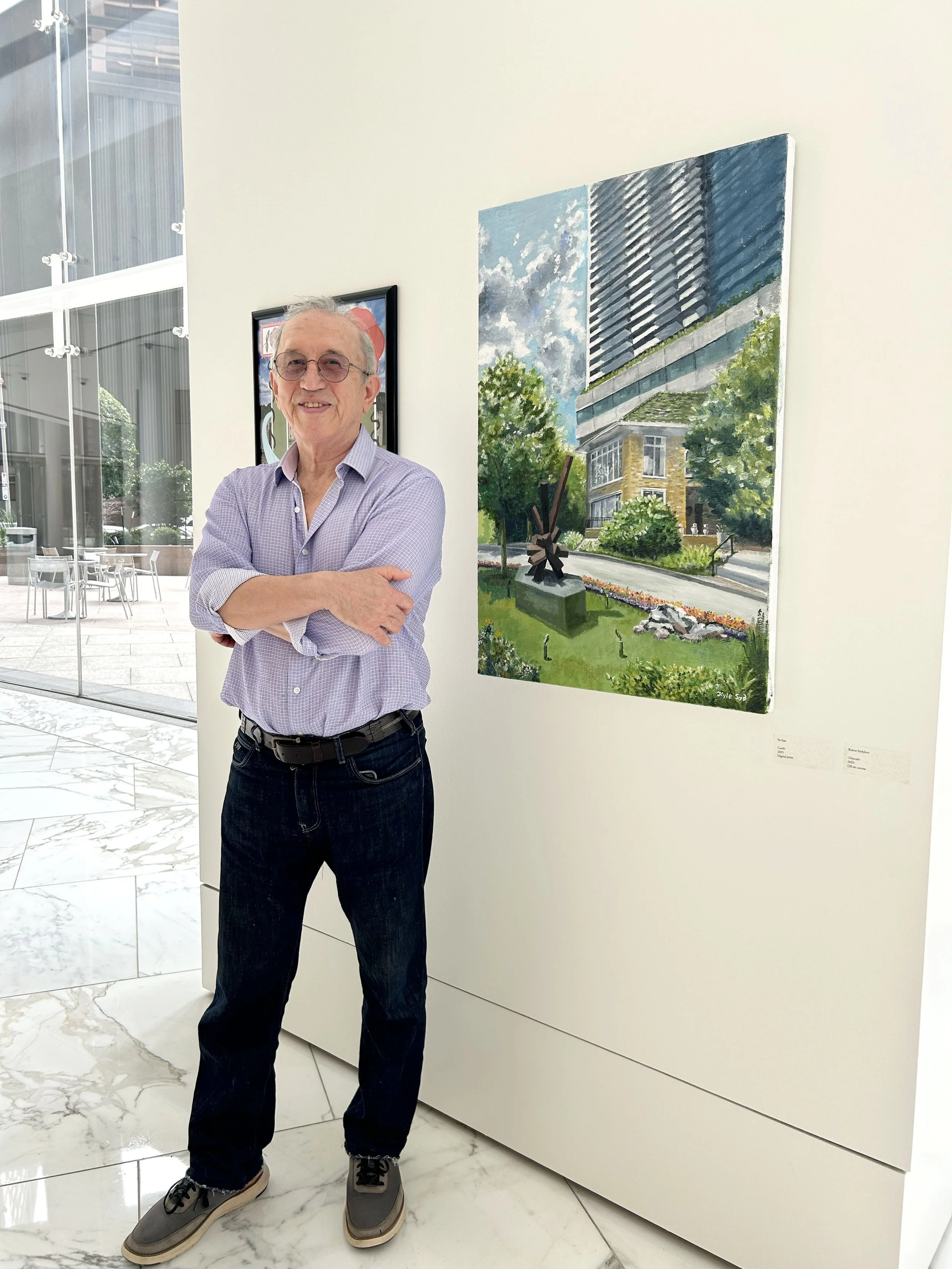 An older man with gray hair and glasses standing with arms crossed in front of a white wall in an art gallery, with a painting of a modern building and trees hanging behind him.