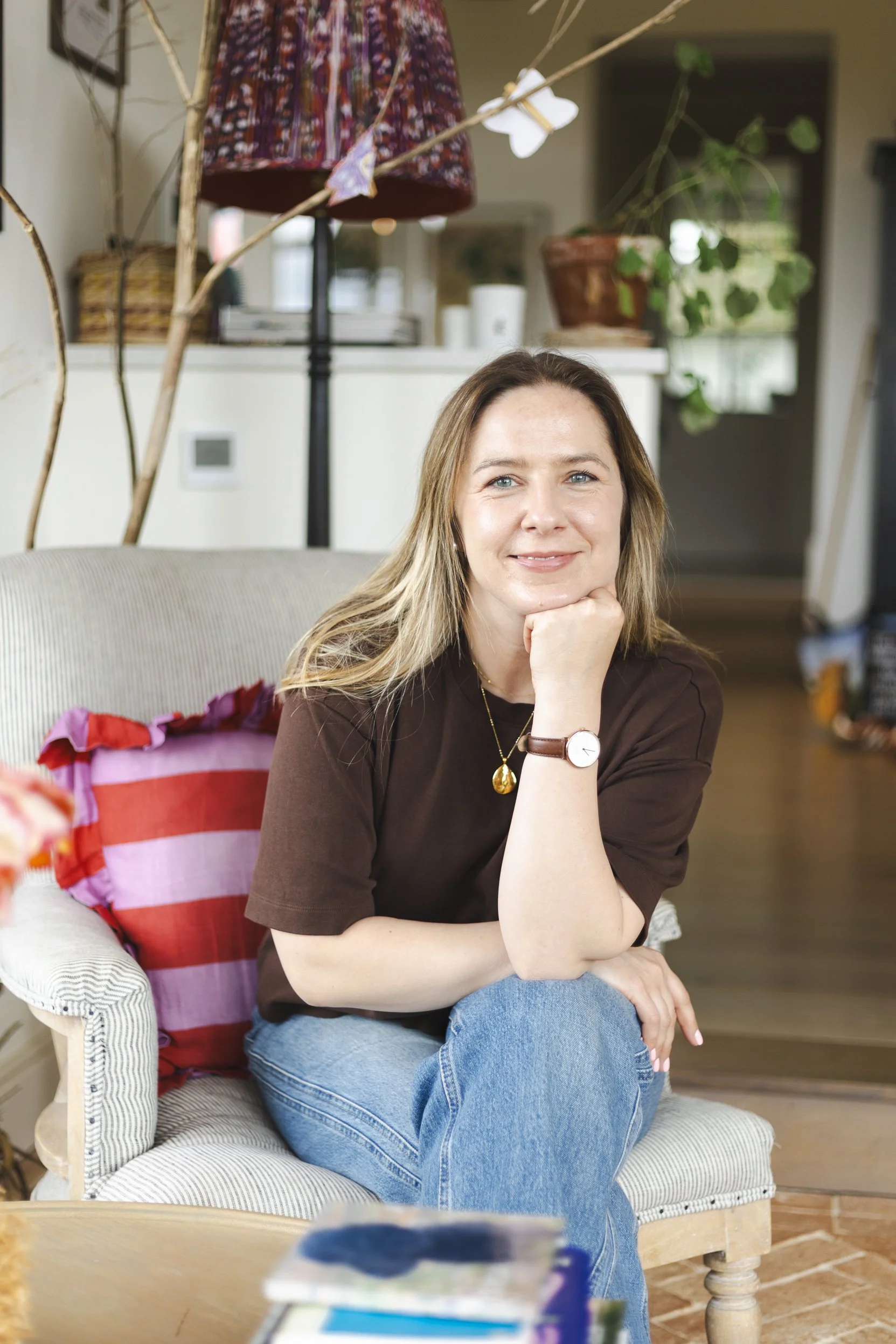 A woman with long light brown hair and blue eyes sits on a beige cushioned armchair with a striped pillow. She is smiling, resting her chin on her right hand, and wearing a dark brown t-shirt, blue jeans, a watch, and a gold necklace. The background shows potted plants, a white shelf with various items, and a hanging tapestry or lamp shade.