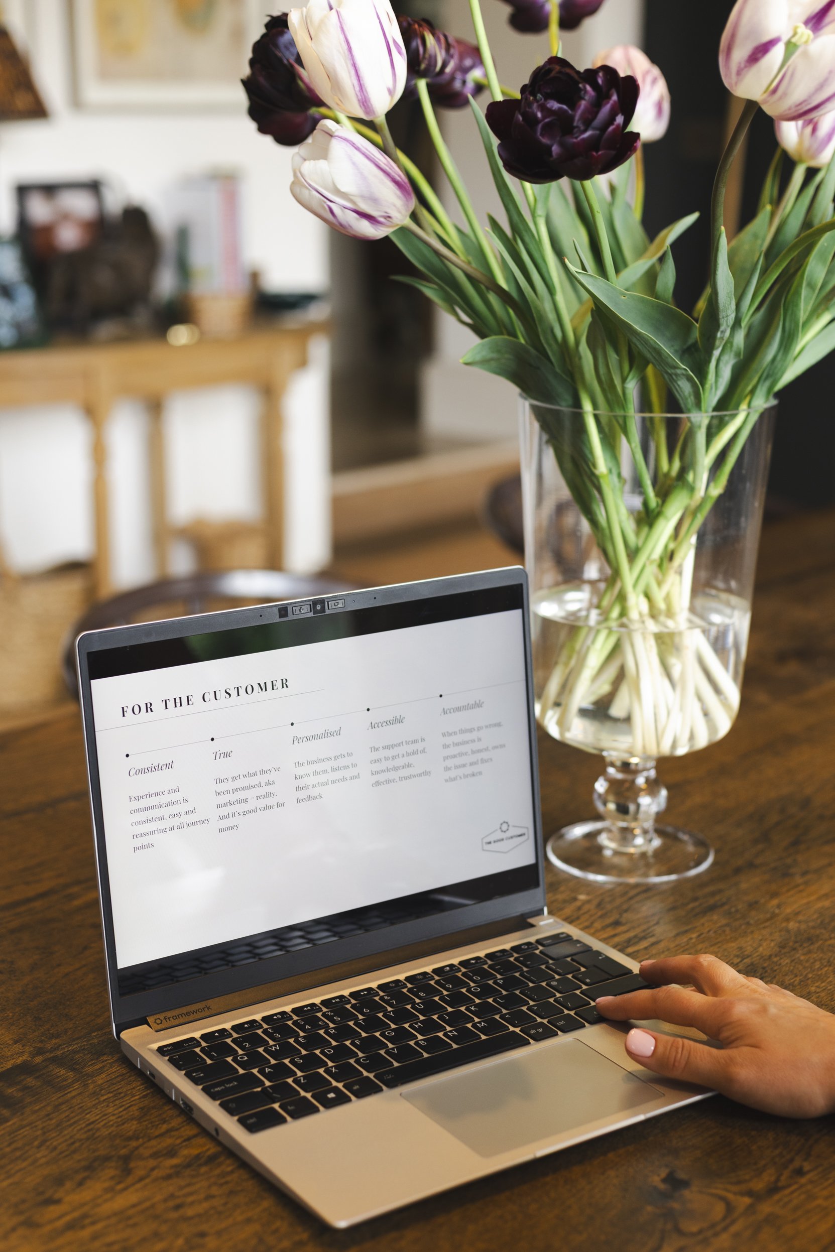 A laptop displaying a document titled 'For the Customer' on a wooden table, with a person's hand near the keyboard. In the background, there is a vase of tulips in various colors, and a blurred indoor setting with furniture.