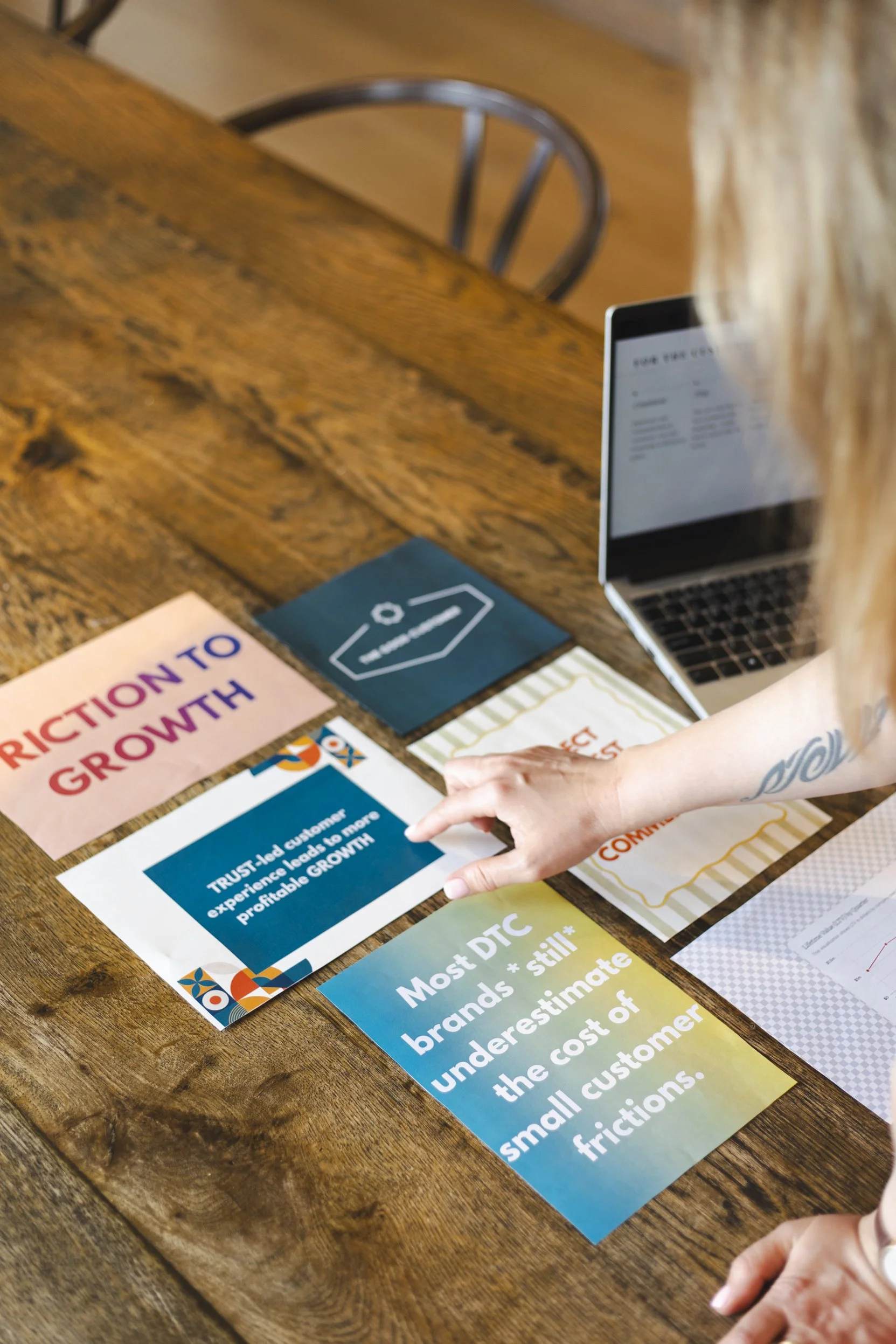 A person's hand pointing at a printed statement on a blue flyer, which is part of a collection of professional brochures and flyers on a wooden table. The person has a tattoo on their forearm.