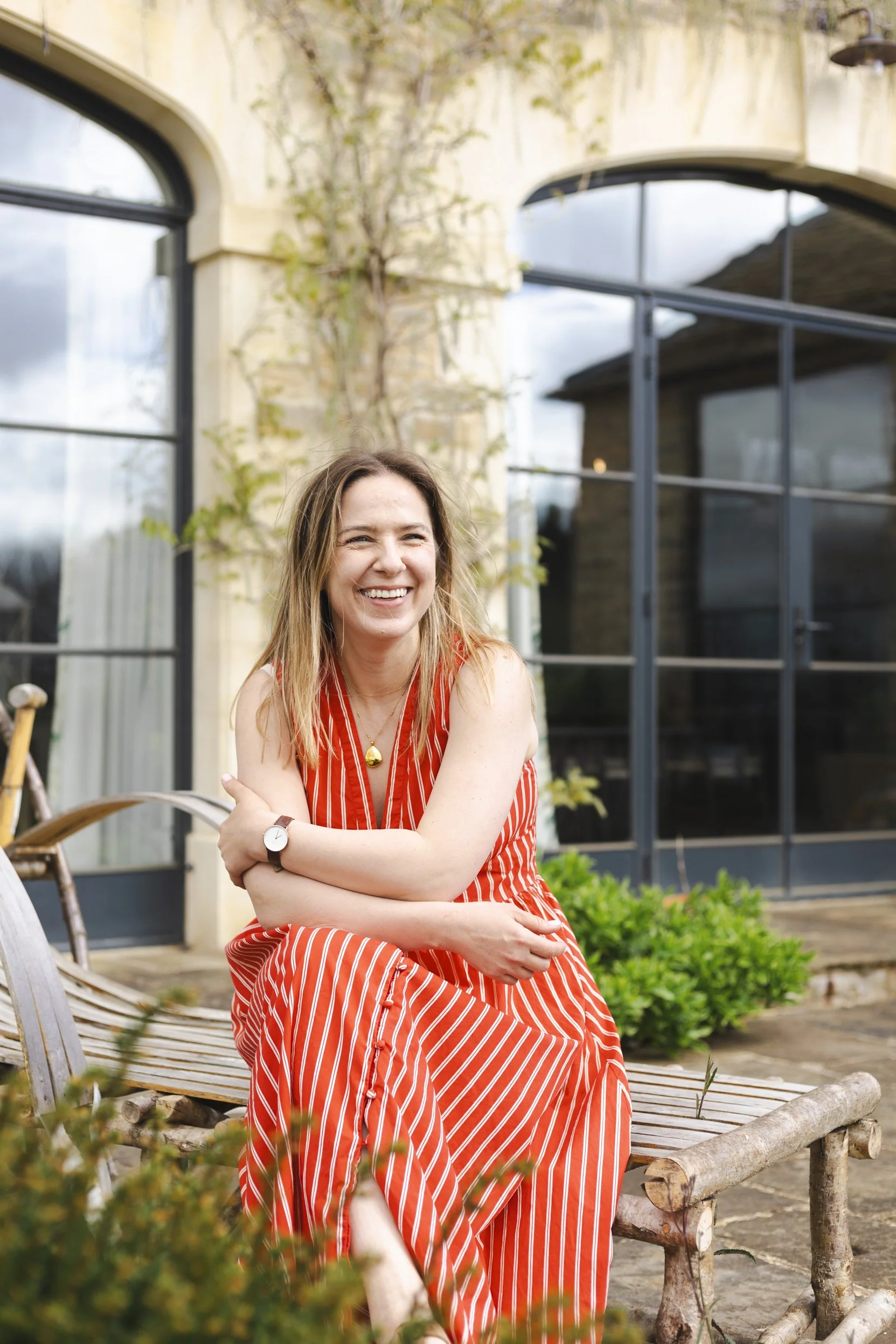 A woman with long hair, wearing a red and white striped dress, sitting outdoors on a wooden bench, smiling with arms crossed, in front of a building with large glass windows and ivy growing on the wall.