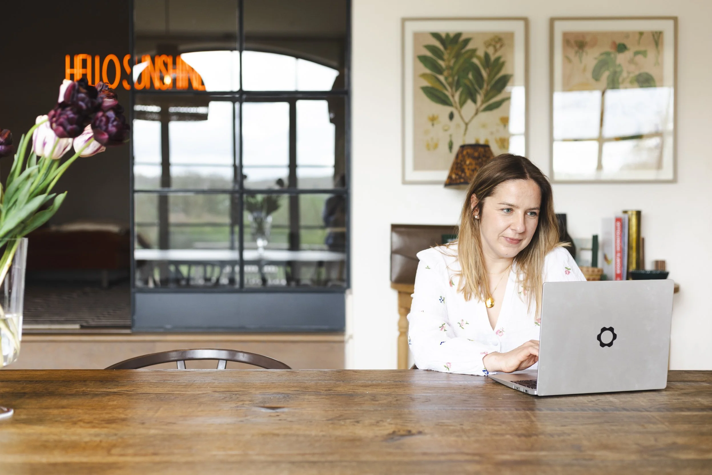A woman with long blonde hair working on a laptop at a wooden dining table in a bright room with artwork on the wall and a large window.