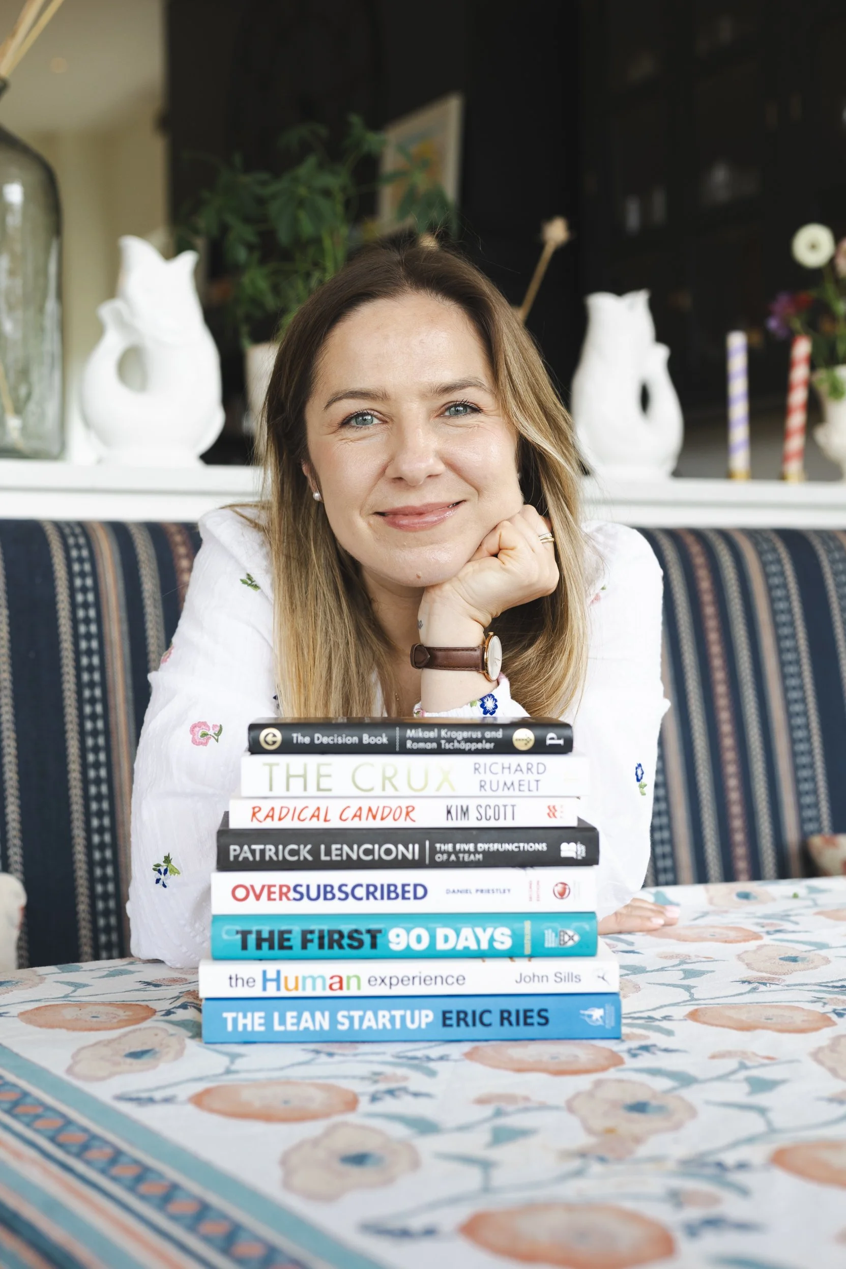 A woman with long light brown hair and blue eyes smiling while resting her chin on her hand, sitting at a table with a stack of books in front of her inside a room decorated with various vases and plants.
