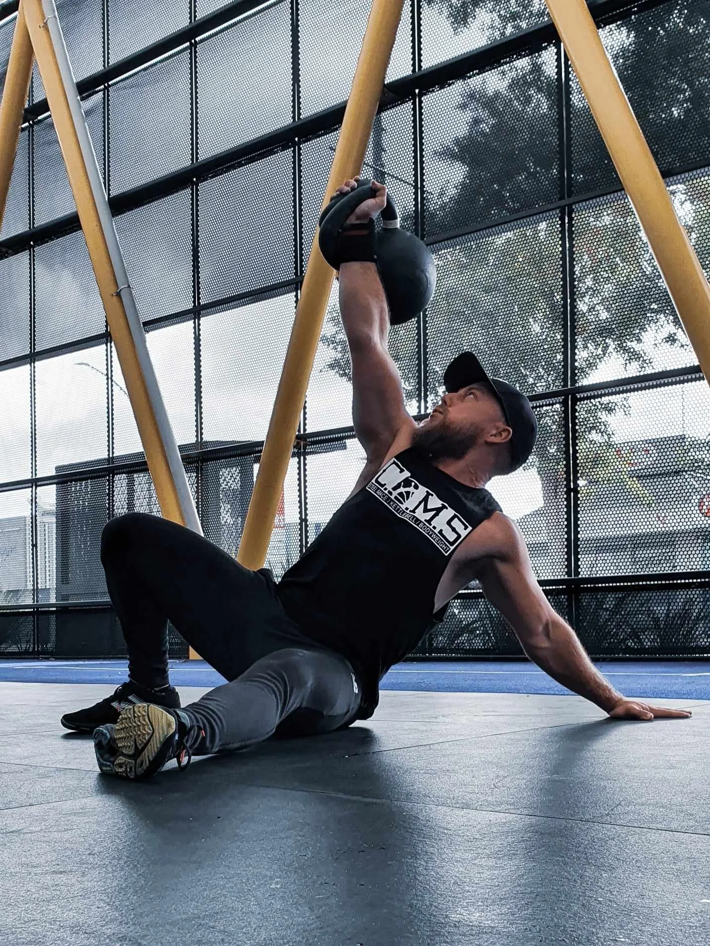 Matthew Selby performing a floor exercise with a kettlebell in a gym with large, black, wire-mesh windows, displaying health and performance