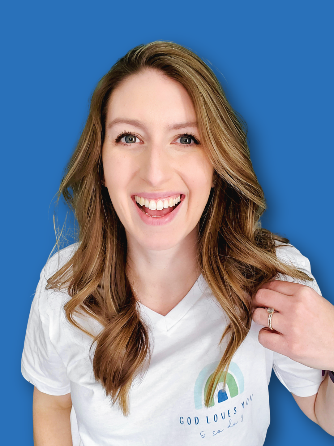 A smiling young woman with wavy, light brown hair against a blue background, wearing a white T-shirt with a colorful rainbow and the text 'GOD LOVES YOU'.