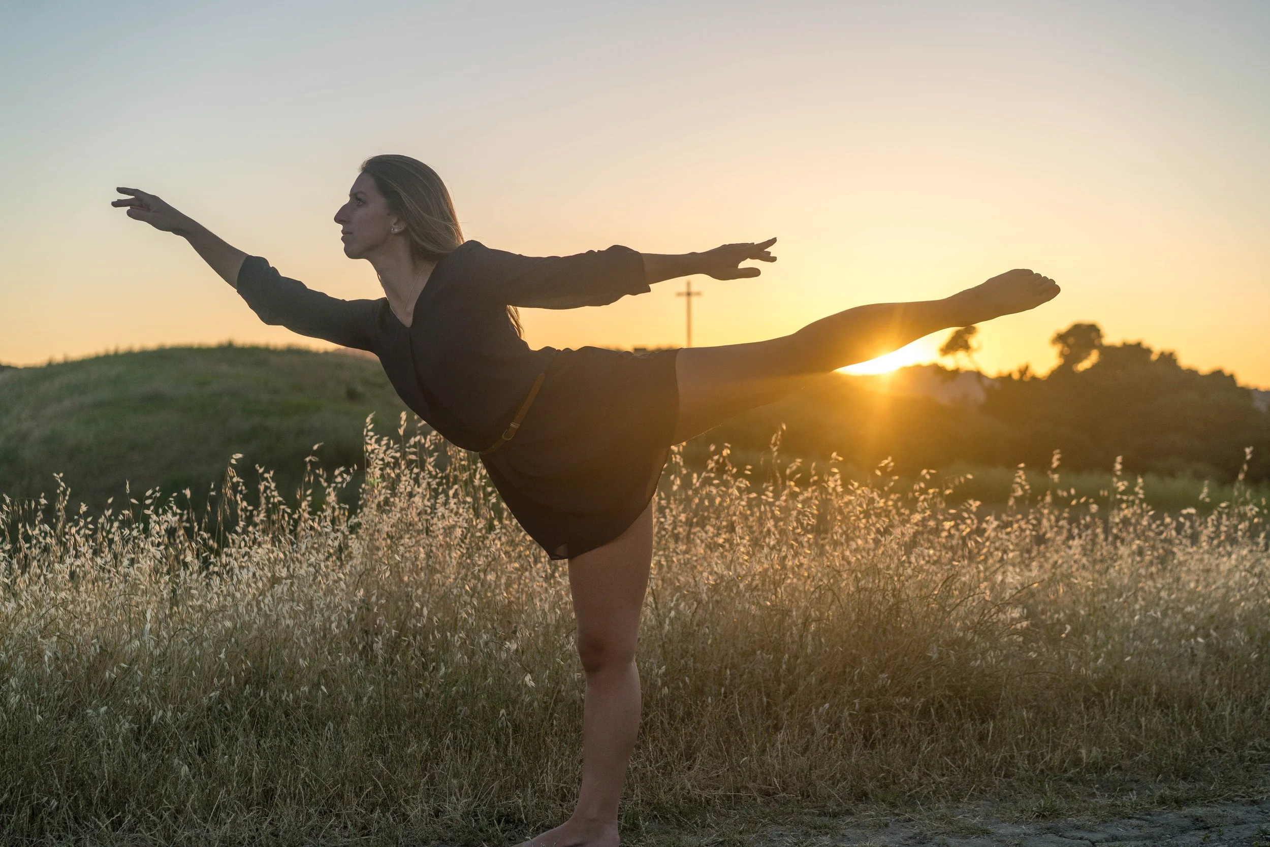 A woman practicing yoga outdoors at sunset, balancing on one leg with arms outstretched, in a grassy field with trees and a cross in the background.