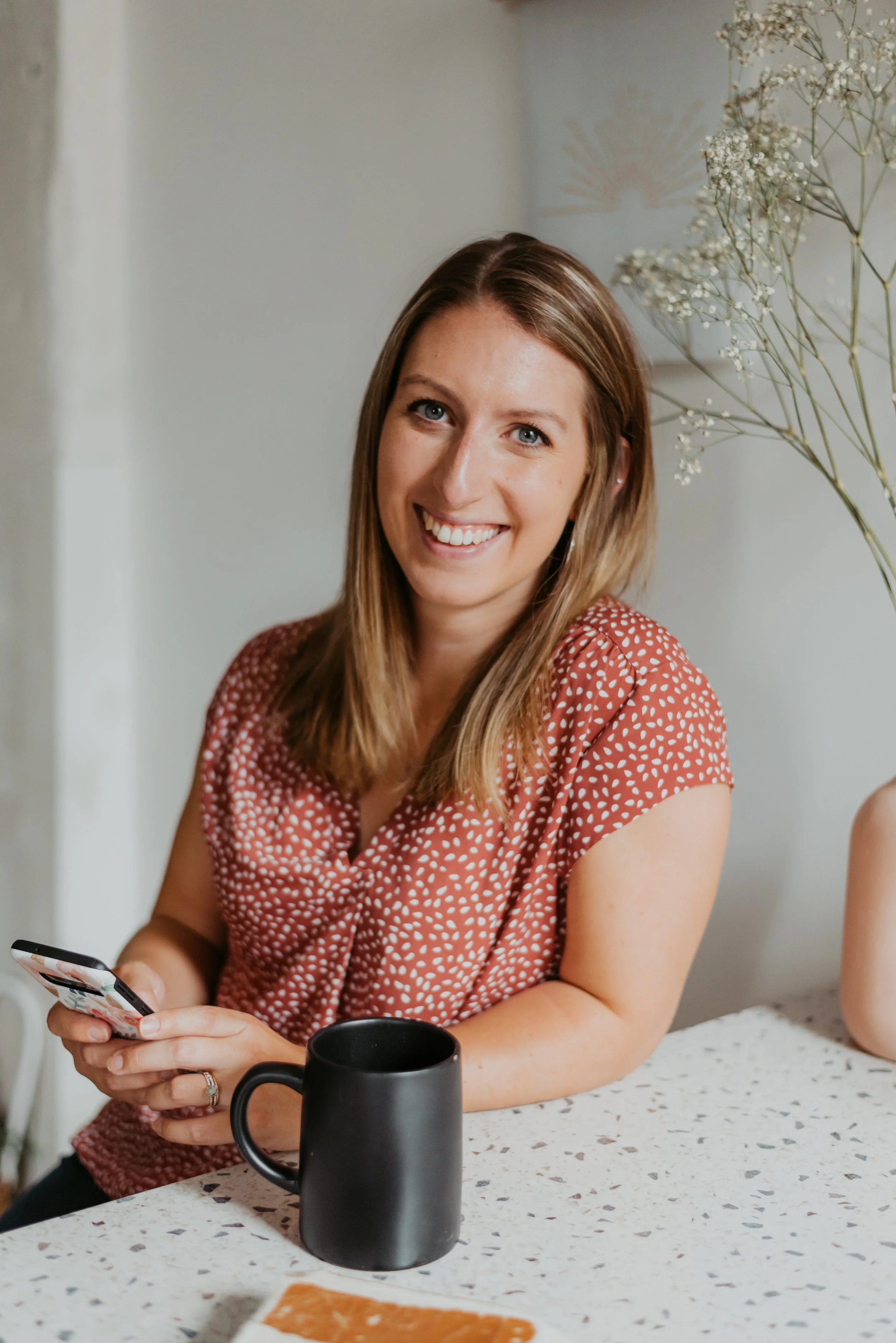 Smiling woman with long blonde hair holding a smartphone, sitting at a speckled countertop with a black mug and a piece of paper, in a bright, casual setting.