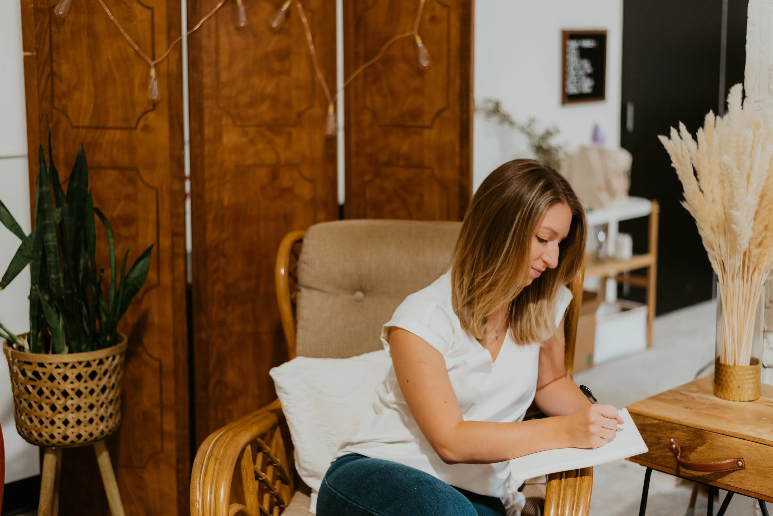 A woman sitting on a beige cushioned chair at a wooden table, writing in a notebook. The setting is a cozy, stylish room with a plant in a woven basket, a wooden screen, decorative dried pampas grass, and other home decor elements.