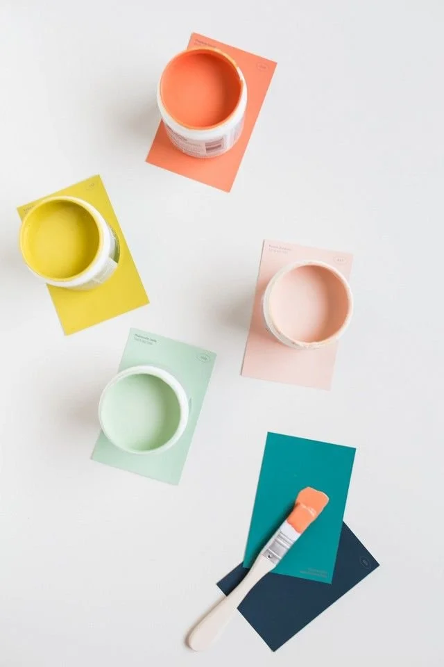Three paint cans in pastel colors (pink, yellow, green) with paint swatches on paper, and a paintbrush with orange paint resting on blue and black paper.