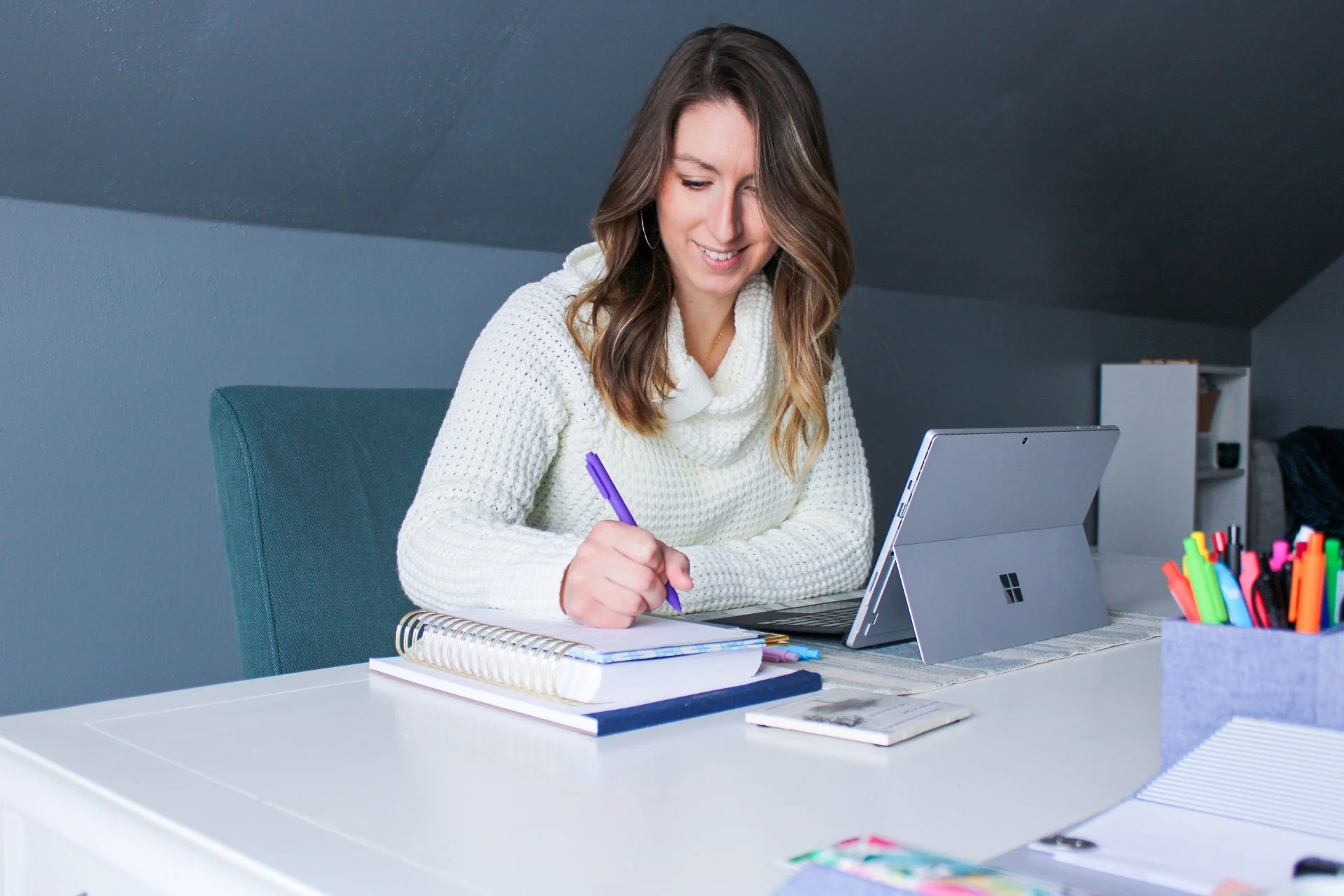 A woman with brown hair, wearing a white sweater, sitting at a white desk with a laptop, notebooks, pens, and markers, writing with a purple pen, smiling while looking at her work.