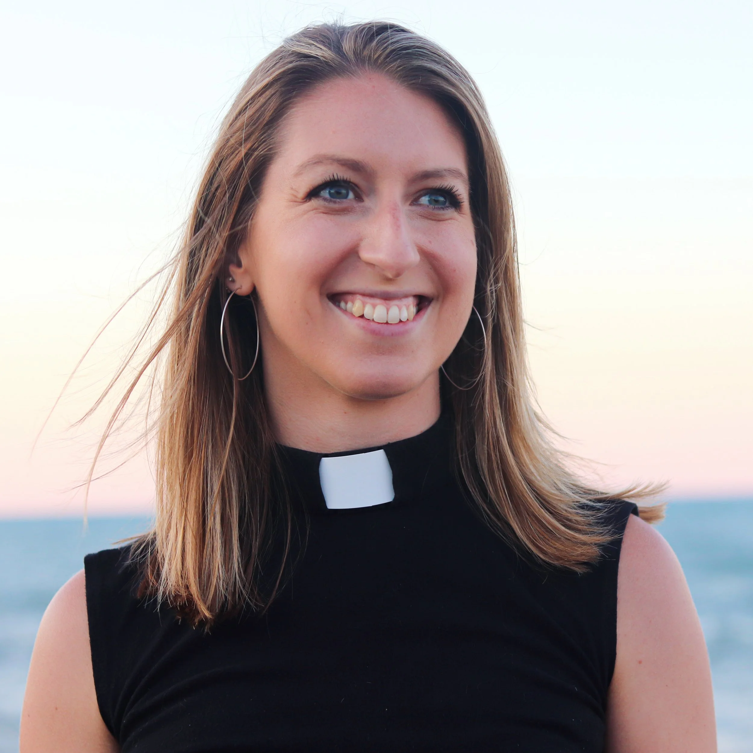 A smiling woman with long brown hair wearing a black clergy shirt and a white clerical collar, standing outdoors near the ocean during sunset.