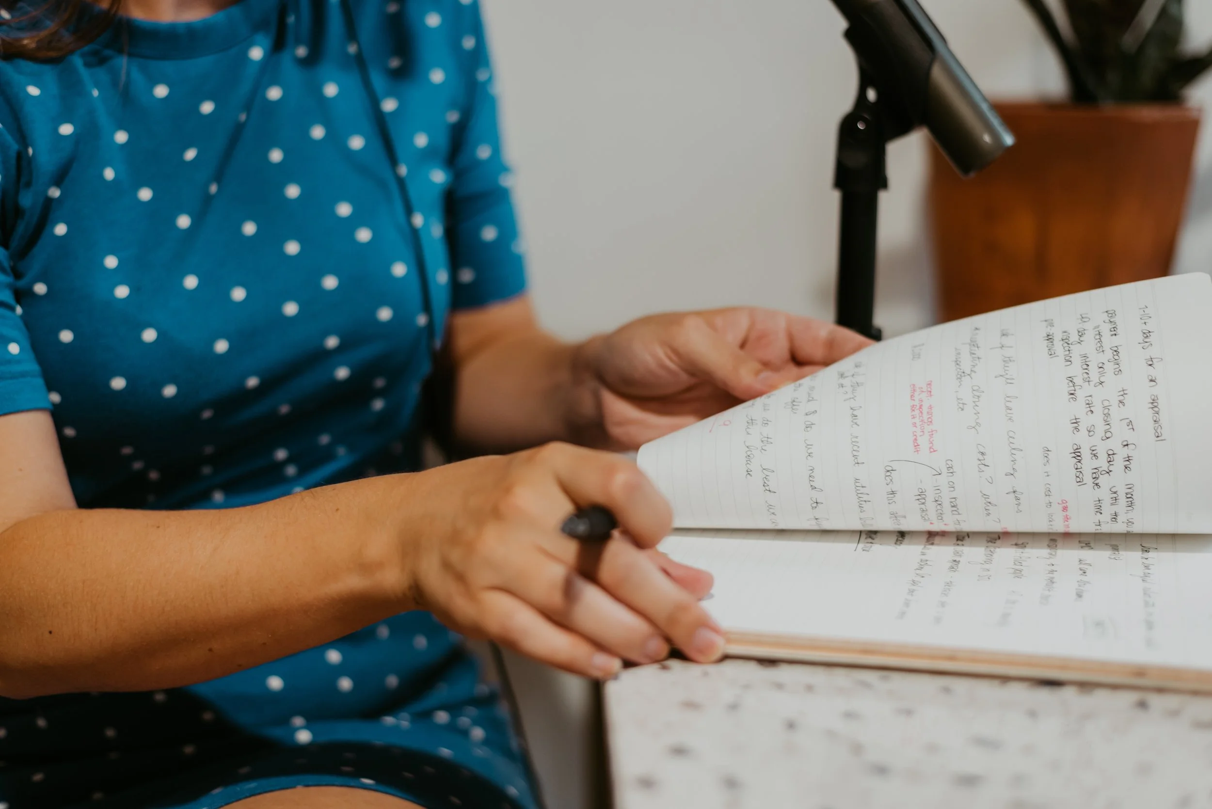 A person wearing a blue polka-dot dress sitting at a table holding a notebook and pen, with a microphone nearby.