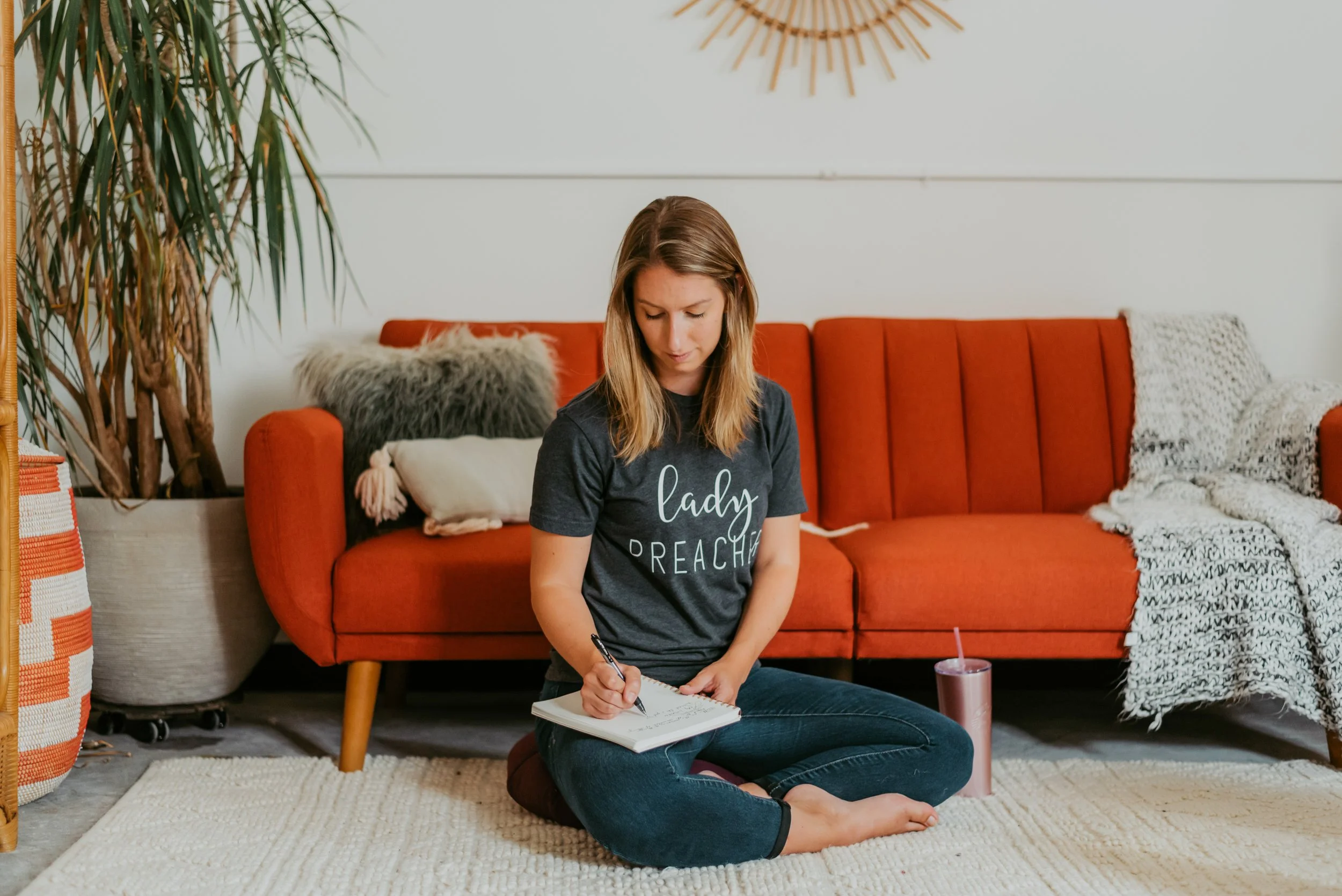 A woman sitting cross-legged on a rug in a living room, writing in a notebook, with a pink tumbler beside her, background features a red sofa with pillows and throws, a large potted plant, and a wall mirror.