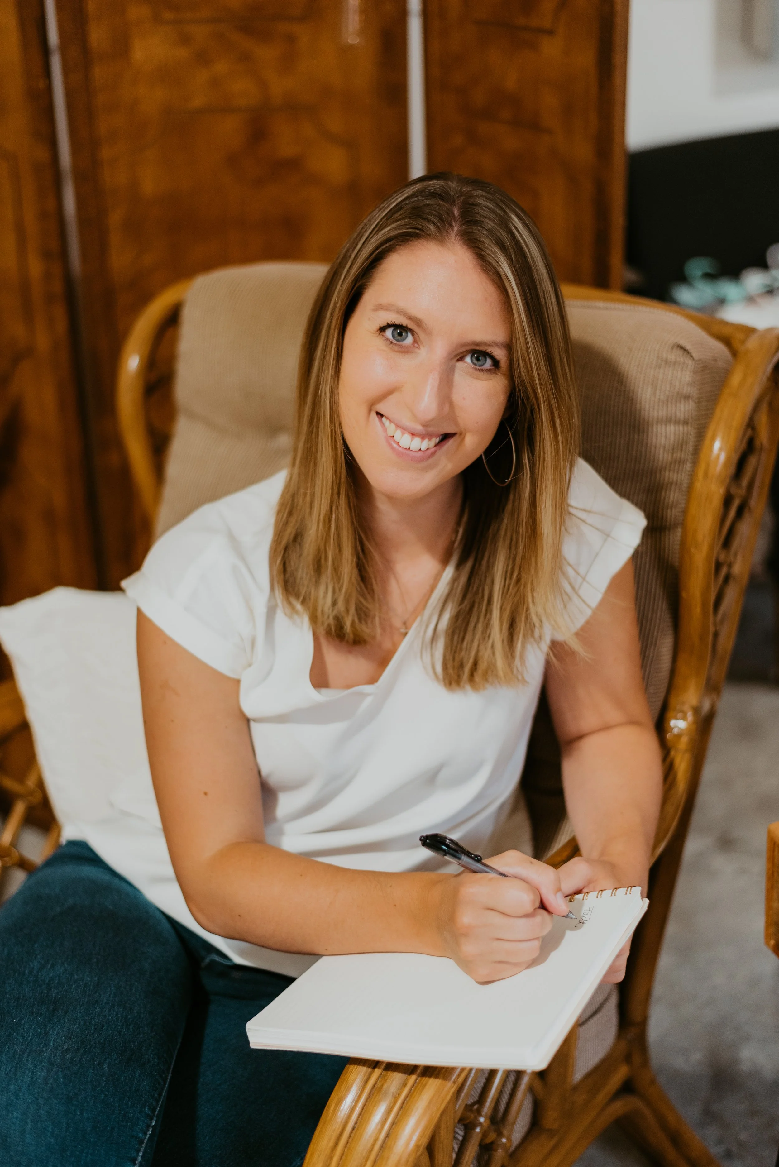 A woman with shoulder-length blonde hair and wearing a white top, sitting on a wooden chair with a cushioned backrest, smiling and holding a black pen while writing in a white notebook.
