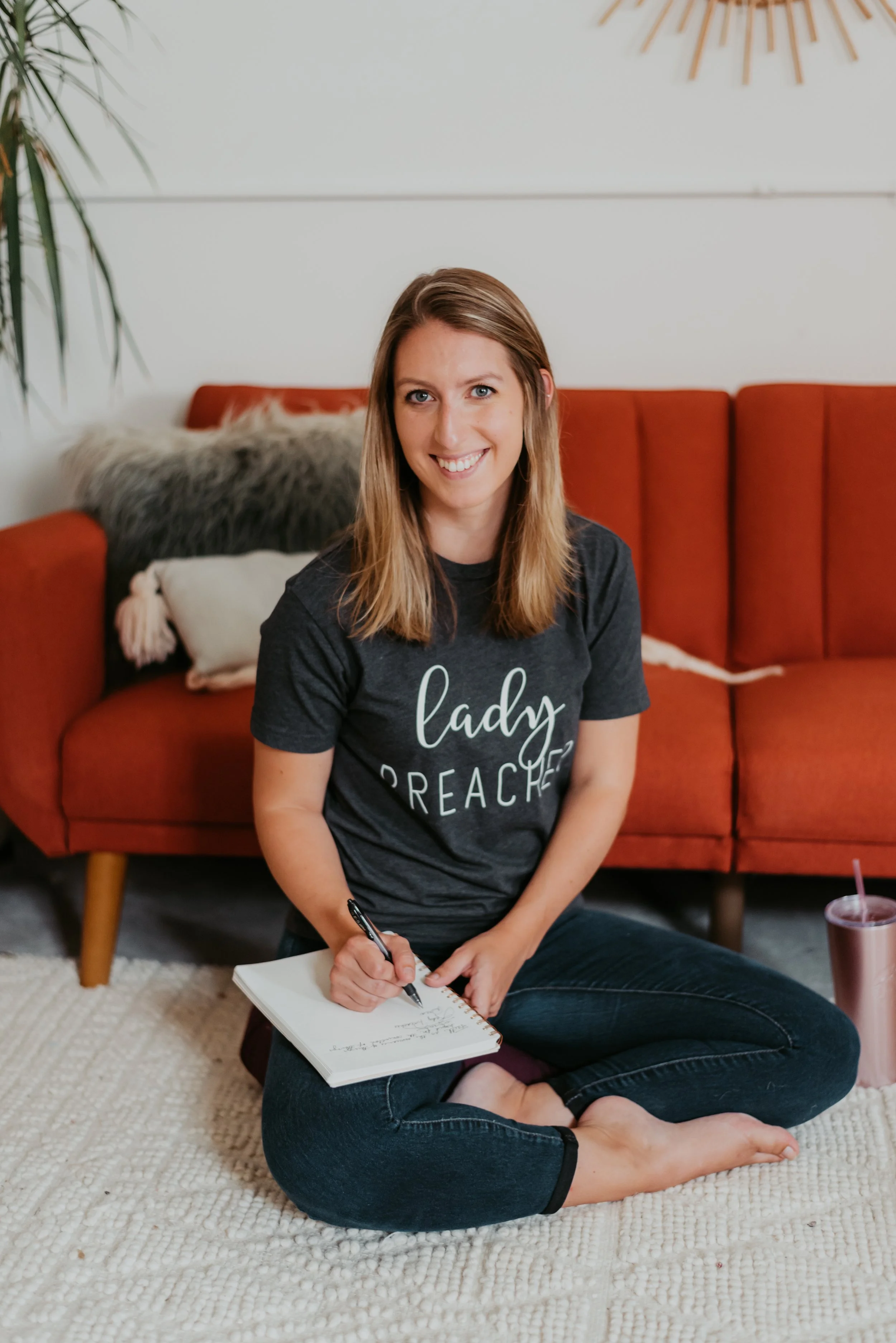 A woman sitting cross-legged on a cream-colored rug in front of a red sofa, smiling and writing in a notebook. She wears a black T-shirt with the words 'lady preacher' and has a tumbler with a straw nearby.