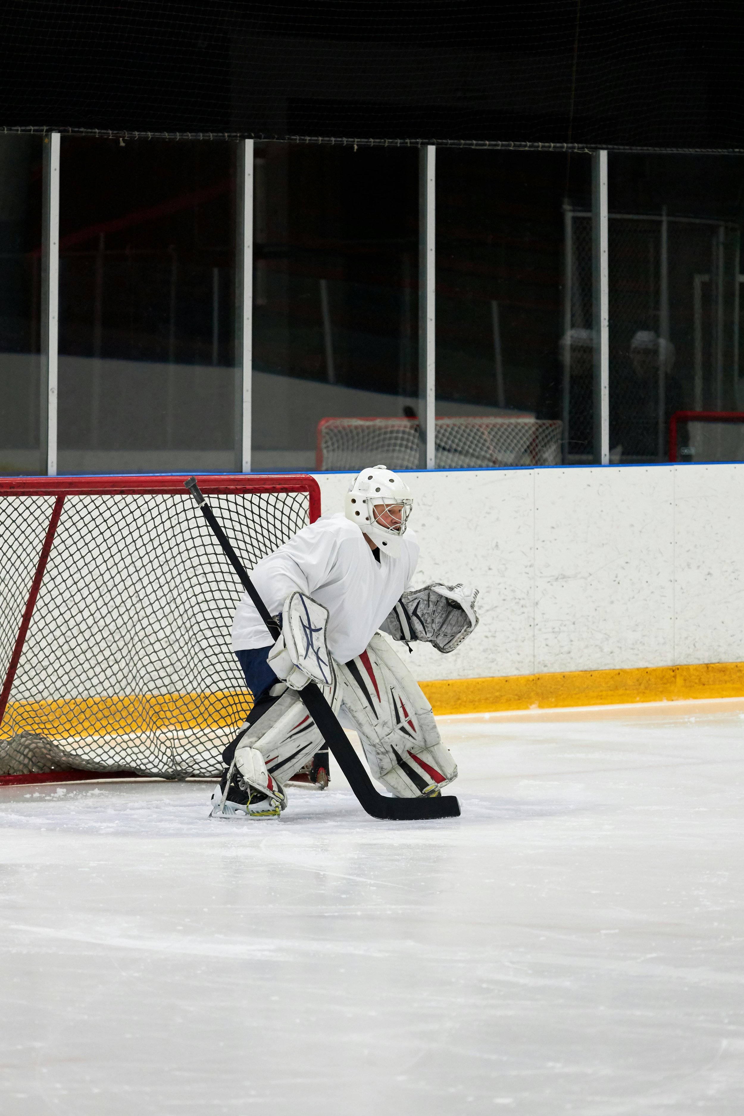Hockey goalie in white gear, crouched in front of goal on ice rink, ready to block a shot.