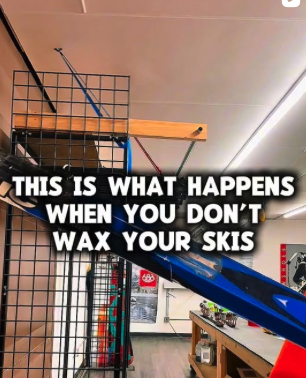 A blue ski leaning against a wooden wall hook inside a store. The background shows a black grid display with other ski poles and accessories, and a wooden countertop with various items. The text overlay reads 'This is what happens when you don't wax your skis.'