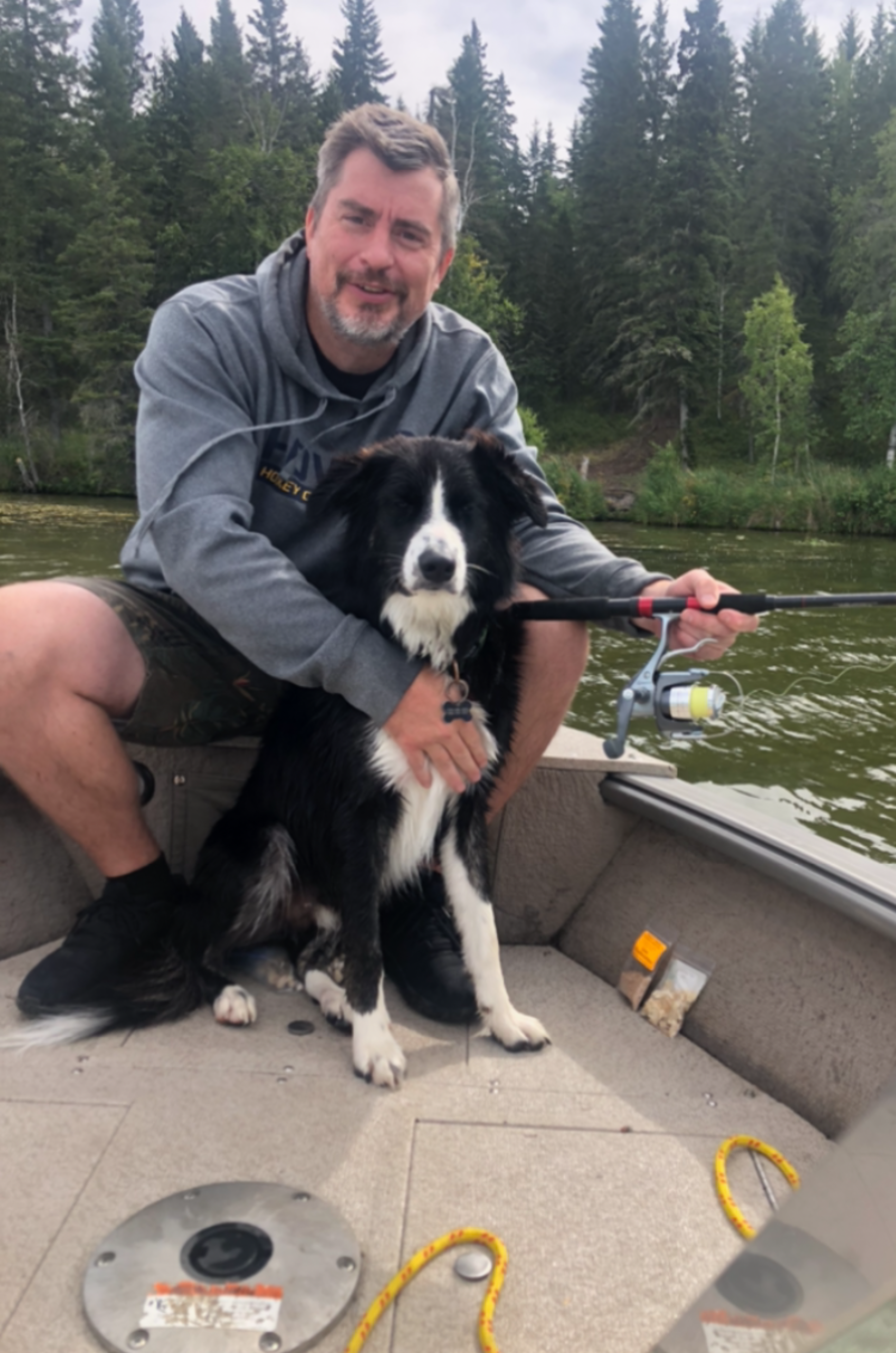 A man in a gray hoodie and shorts is sitting in a boat, holding a fishing rod and a black and white Border Collie sitting beside him. They are on a body of water with a forested shoreline in the background.