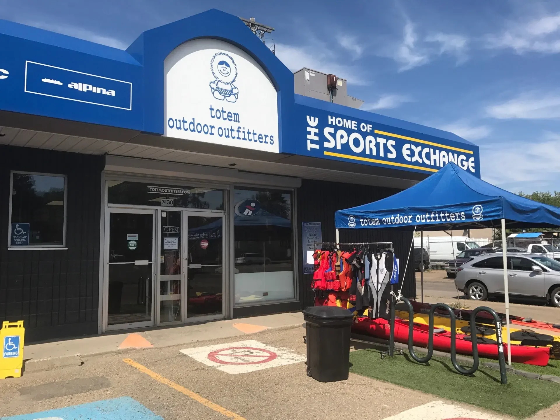 Exterior view of Totem Outdoor Outfitters store with outdoor gear, kayaks, and lif jackets displayed outside under a blue canopy. Signage includes 'The Home of the Sports Exchange' and 'Totem Outdoor Outfitters.' Parking lot with cars and handicap parking signs visible.