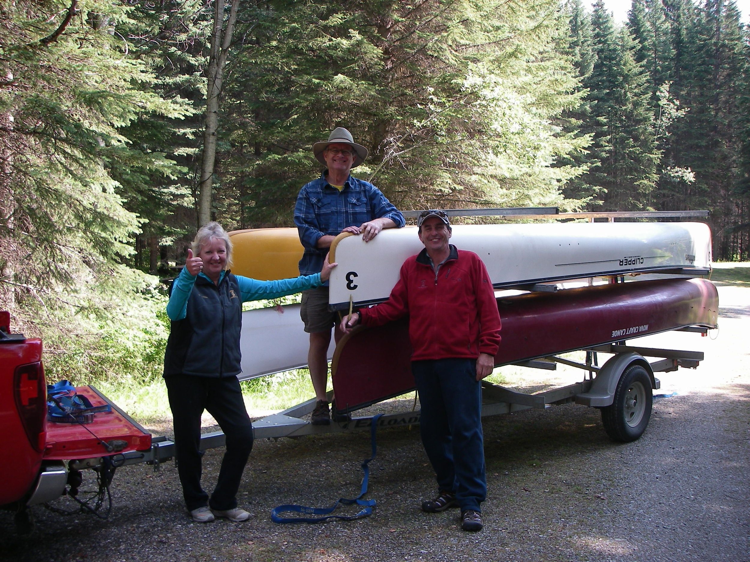 Three people with a canoe and kayak on a trailer in a forested area. One person is giving a thumbs up. All are smiling and dressed in outdoor gear.