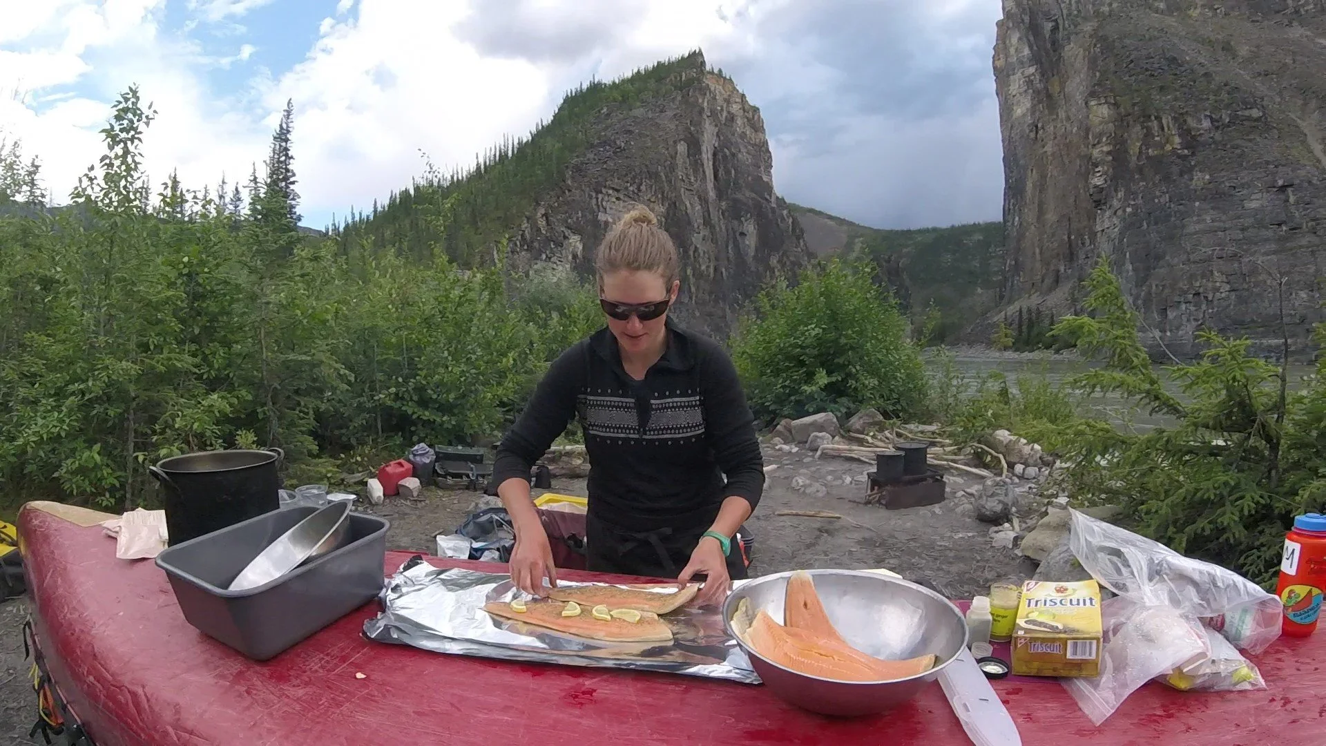 A woman with sunglasses and a ponytail preparing food outdoors at a camping site with mountains, trees, and a river in the background.